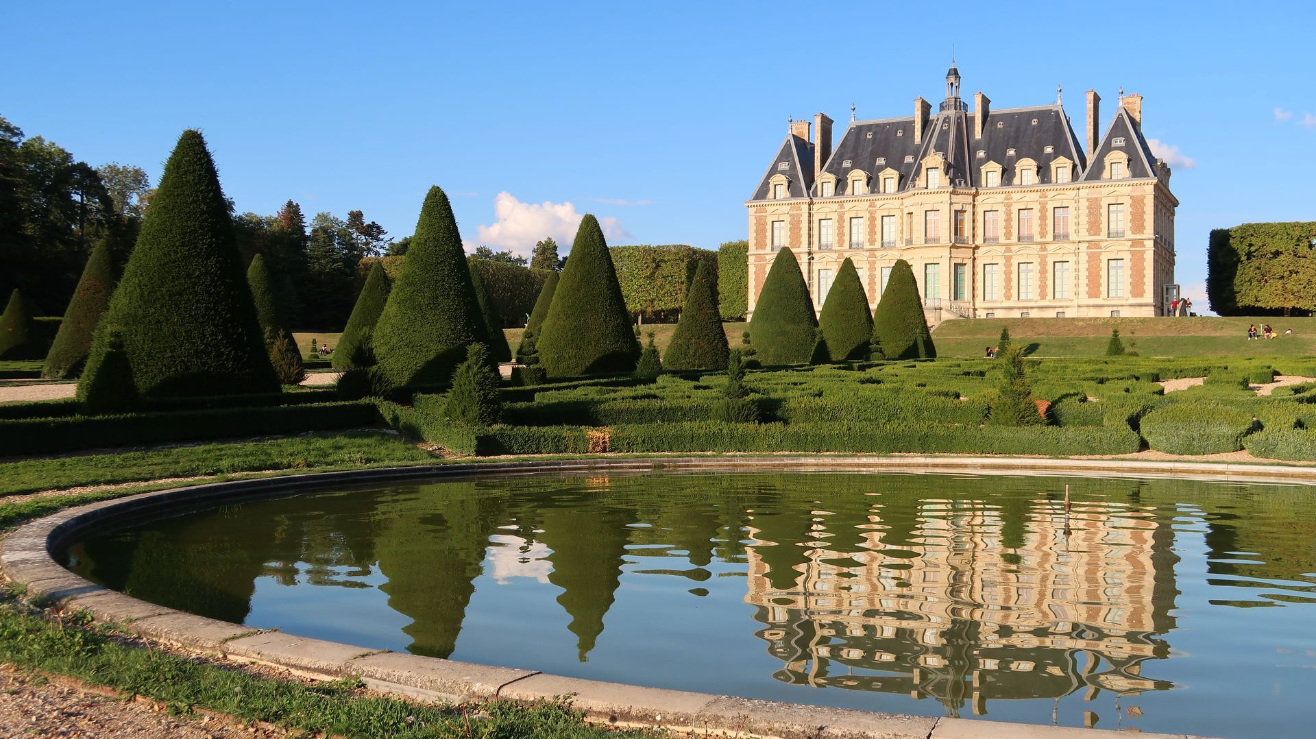 Un grand château au toit à forte pente se reflète dans un bassin de jardin à la française, entouré de haies coniques sous un ciel bleu.