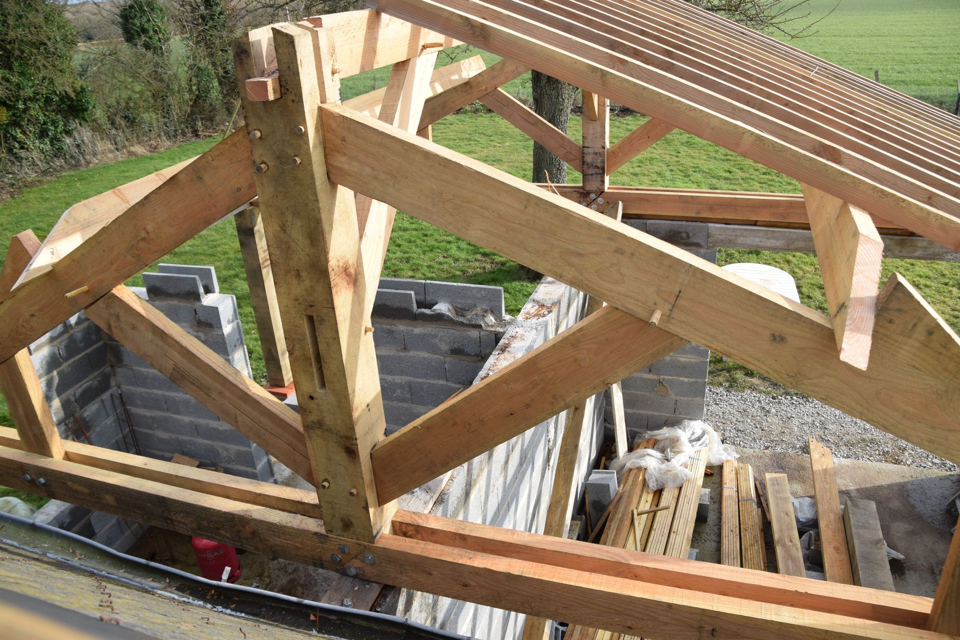 Charpente de toit en bois en construction pour un bâtiment extérieur avec vue sur un champ herbeux en arrière-plan.