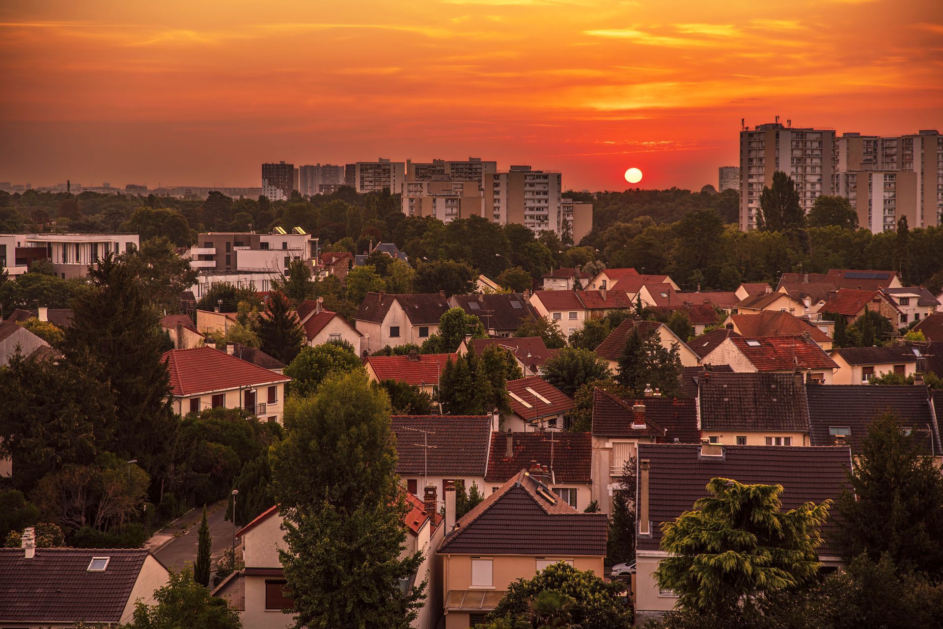 Au coucher du soleil, une lueur orangée illumine un quartier résidentiel où se mêlent maisons et immeubles d'appartements plus hauts, au milieu d'arbres verdoyants.