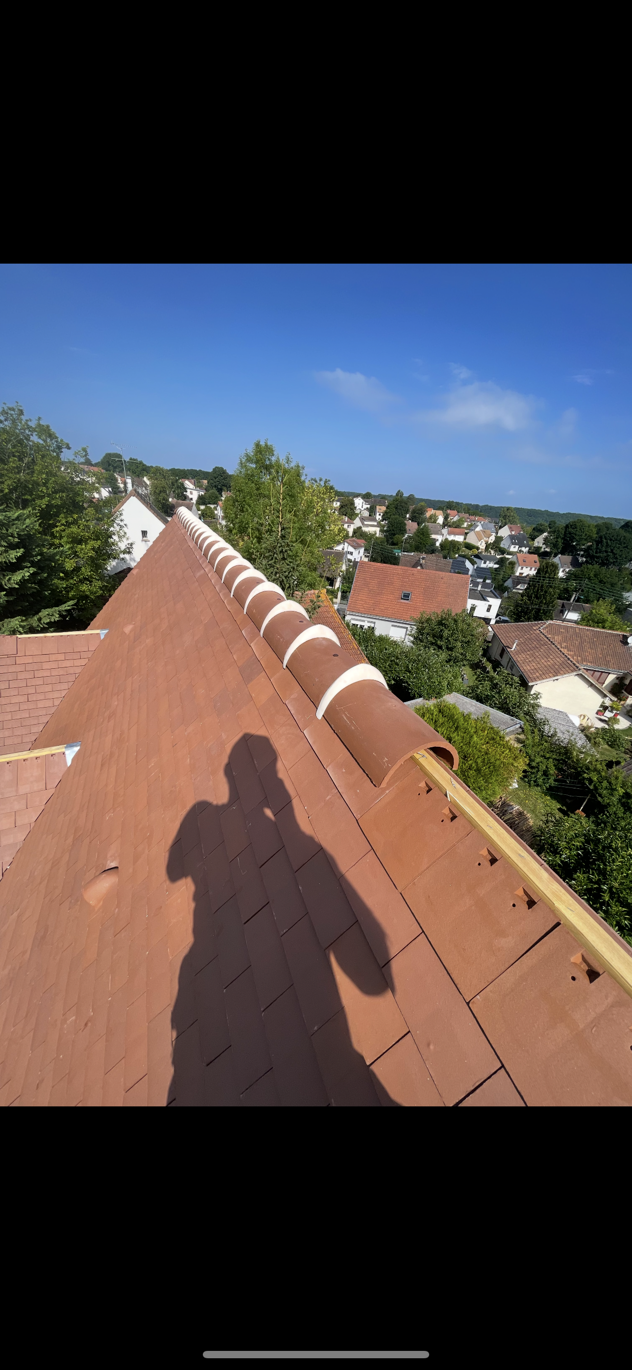 Vue depuis un toit montrant l'ombre d'une personne sur des bardeaux rouges près d'une crête, surplombant une ville par une journée ensoleillée.