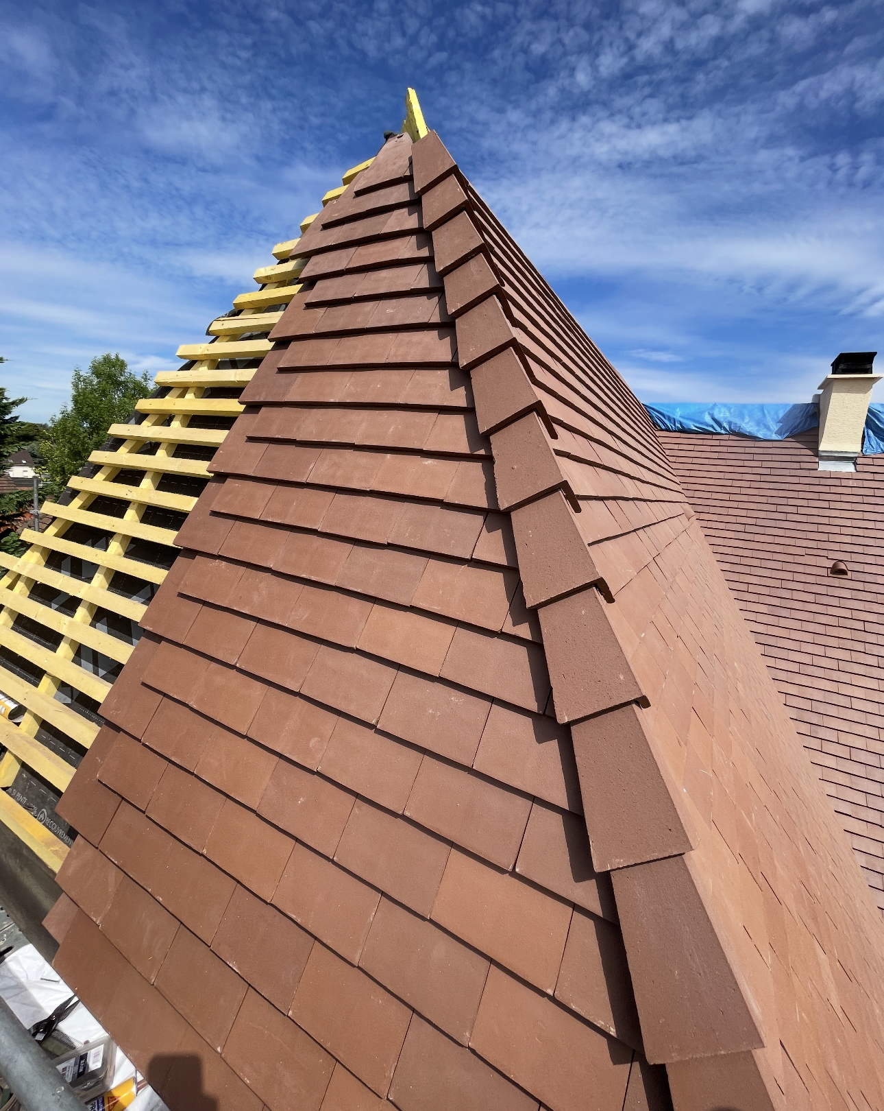 Un toit pentu en construction, recouvert de tuiles en terre cuite brune, avec une charpente en bois apparente, et un ciel d'un bleu limpide.