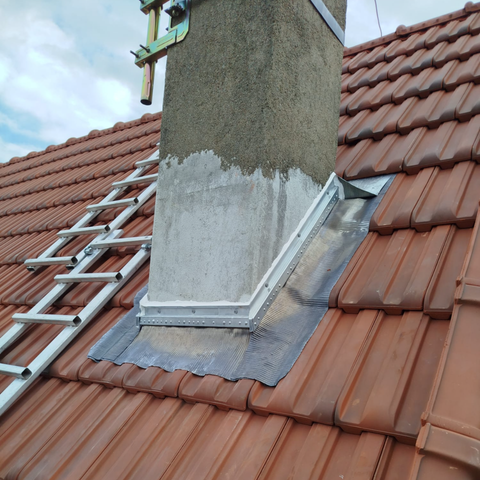 Une cheminée sur un toit de tuiles avec des solins gris neufs, du mastic d'étanchéité et une échelle métallique appuyée contre le côté.