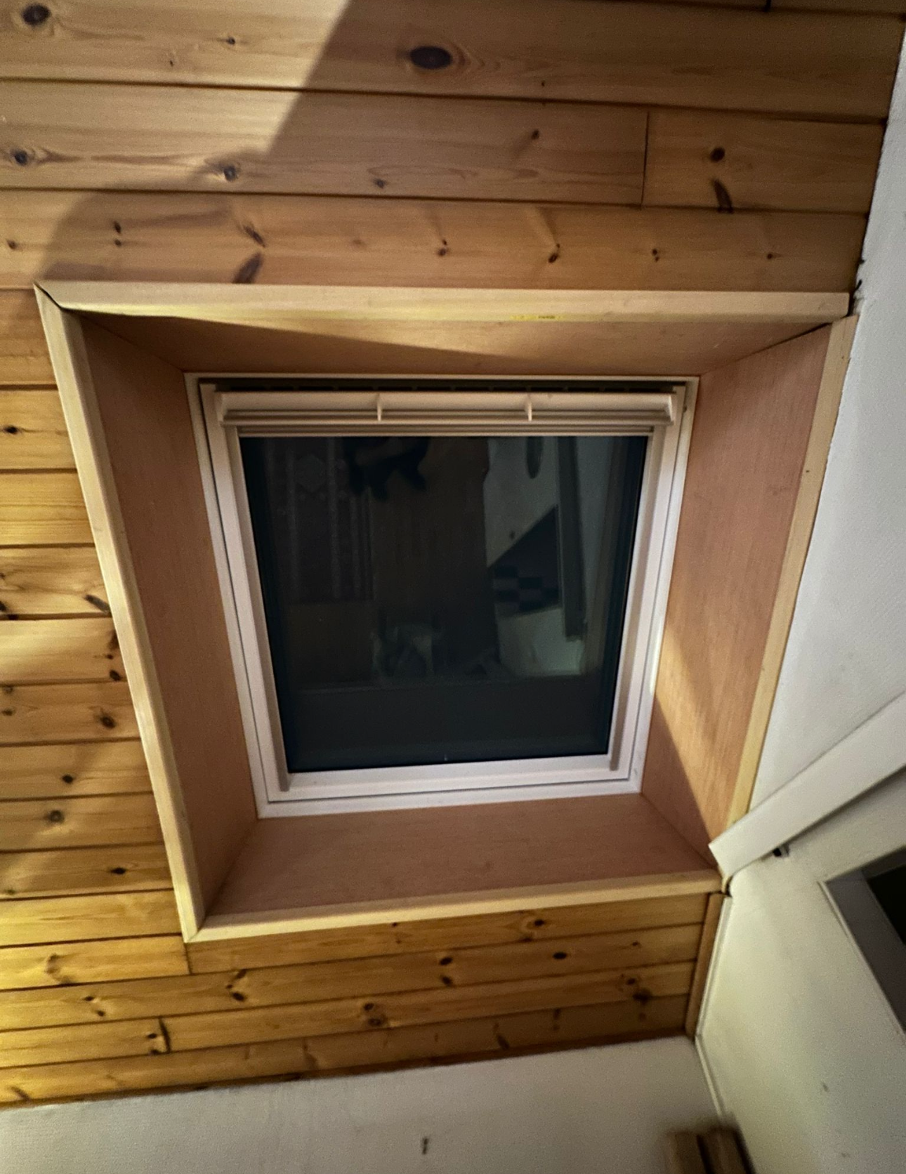 Une fenêtre de toit carrée encadrée de panneaux de bois clair non finis, intégrée à un mur de planches de bois horizontales.