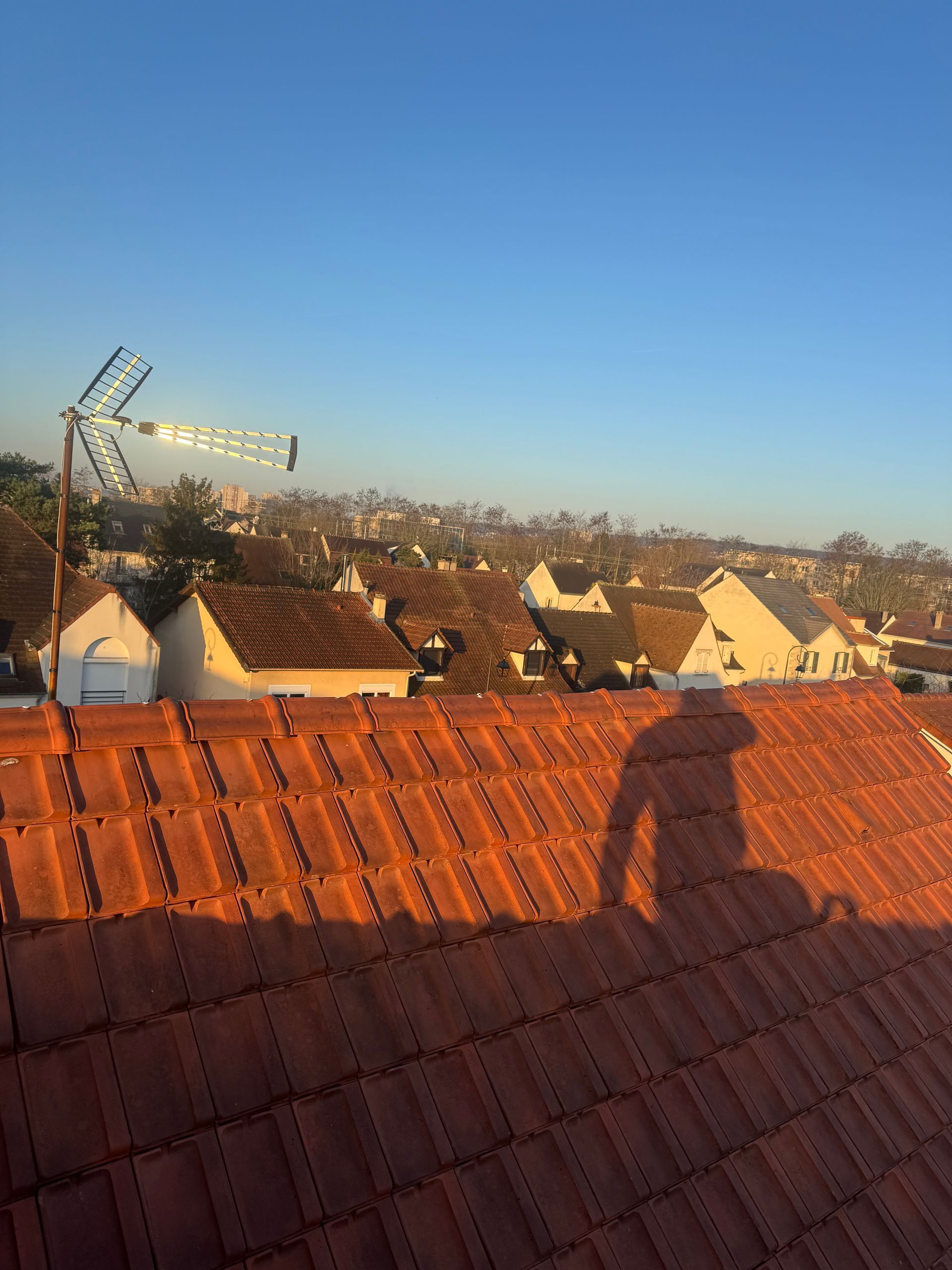 Vue depuis un toit de tuiles donnant sur un quartier au coucher du soleil, avec une antenne de télévision et une ombre projetée sur le toit.