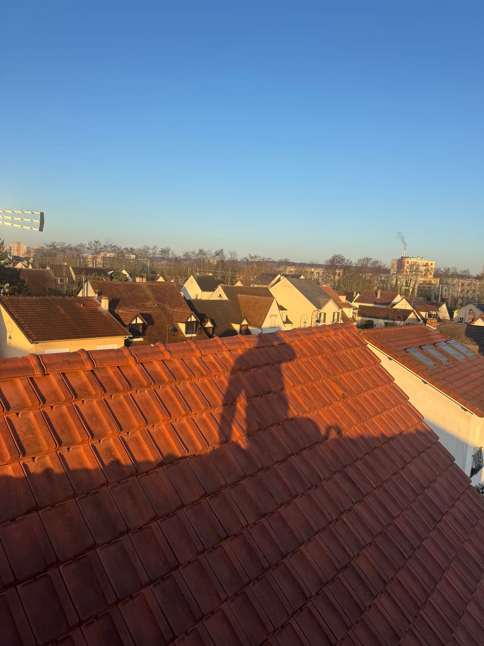 L'ombre d'une personne se projette sur un toit de tuiles, avec un ciel bleu clair et un paysage urbain en arrière-plan.