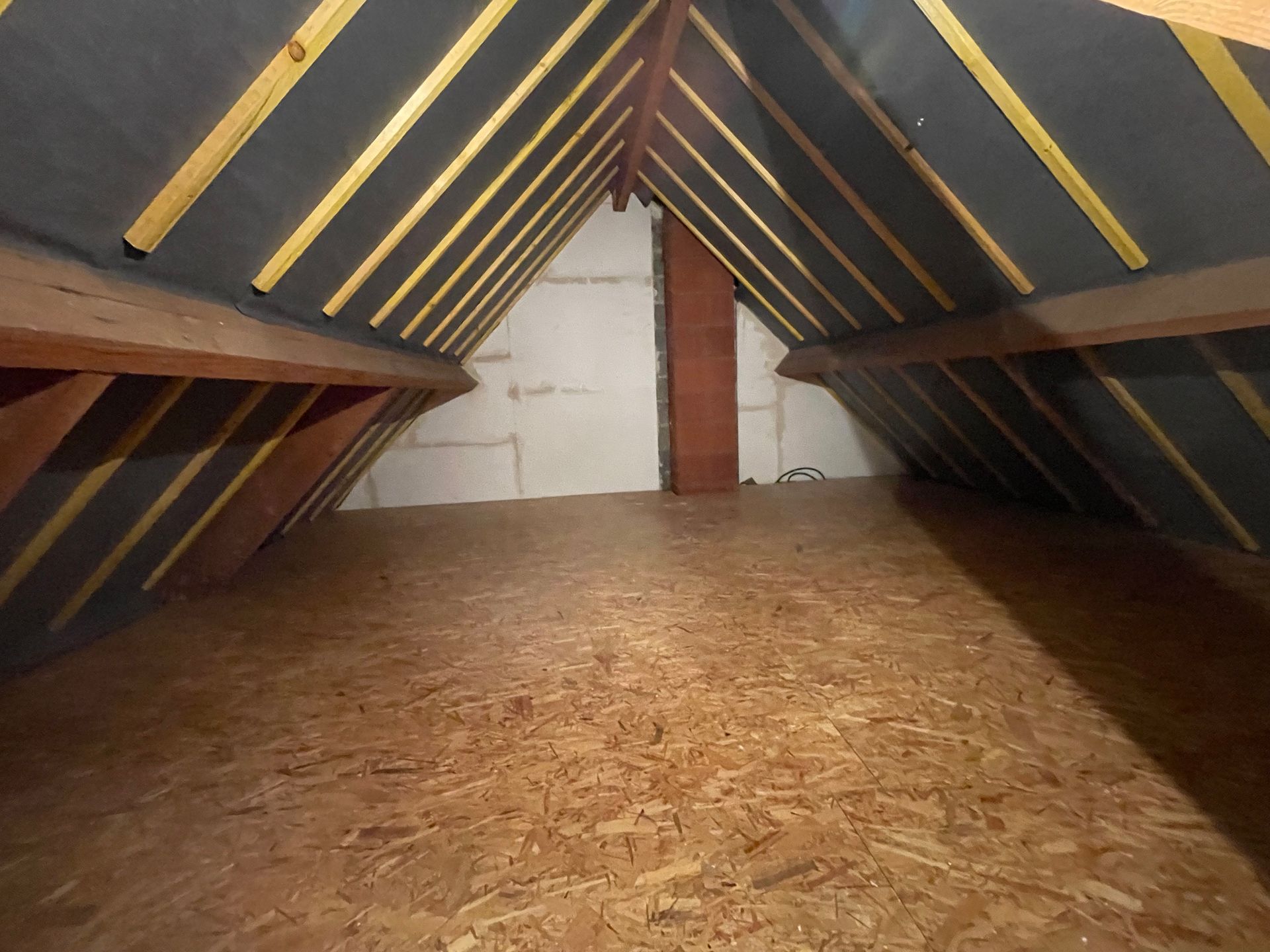 Un grenier avec une charpente en bois, une isolation sombre sous la toiture, une cheminée en briques brunes et un plancher en contreplaqué.