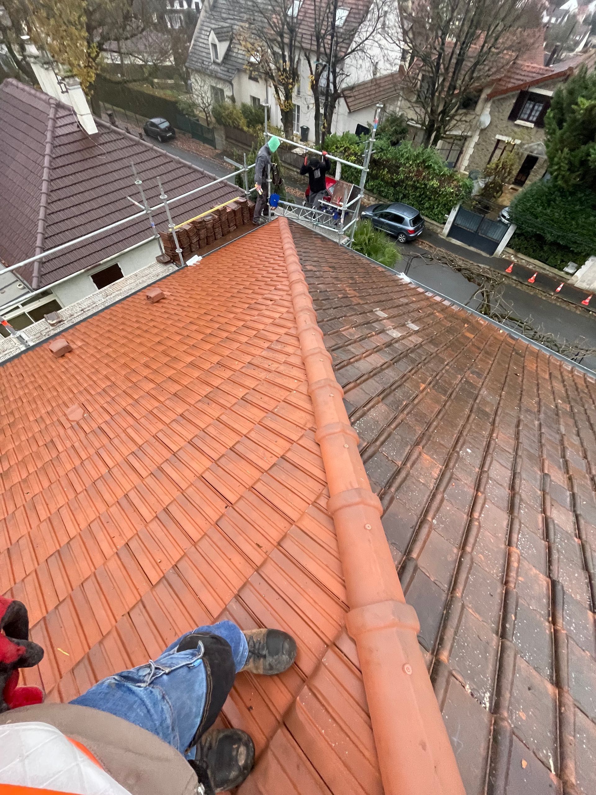 Un ouvrier vêtu d'un équipement haute visibilité se tient sur un toit de tuiles brun-rougeâtre partiellement rénové, donnant sur une rue tranquille.