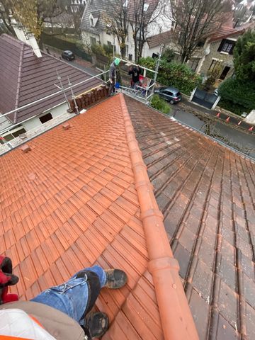 Un ouvrier vêtu d'un équipement haute visibilité se tient sur un toit de tuiles brun-rougeâtre partiellement rénové, donnant sur une rue tranquille.