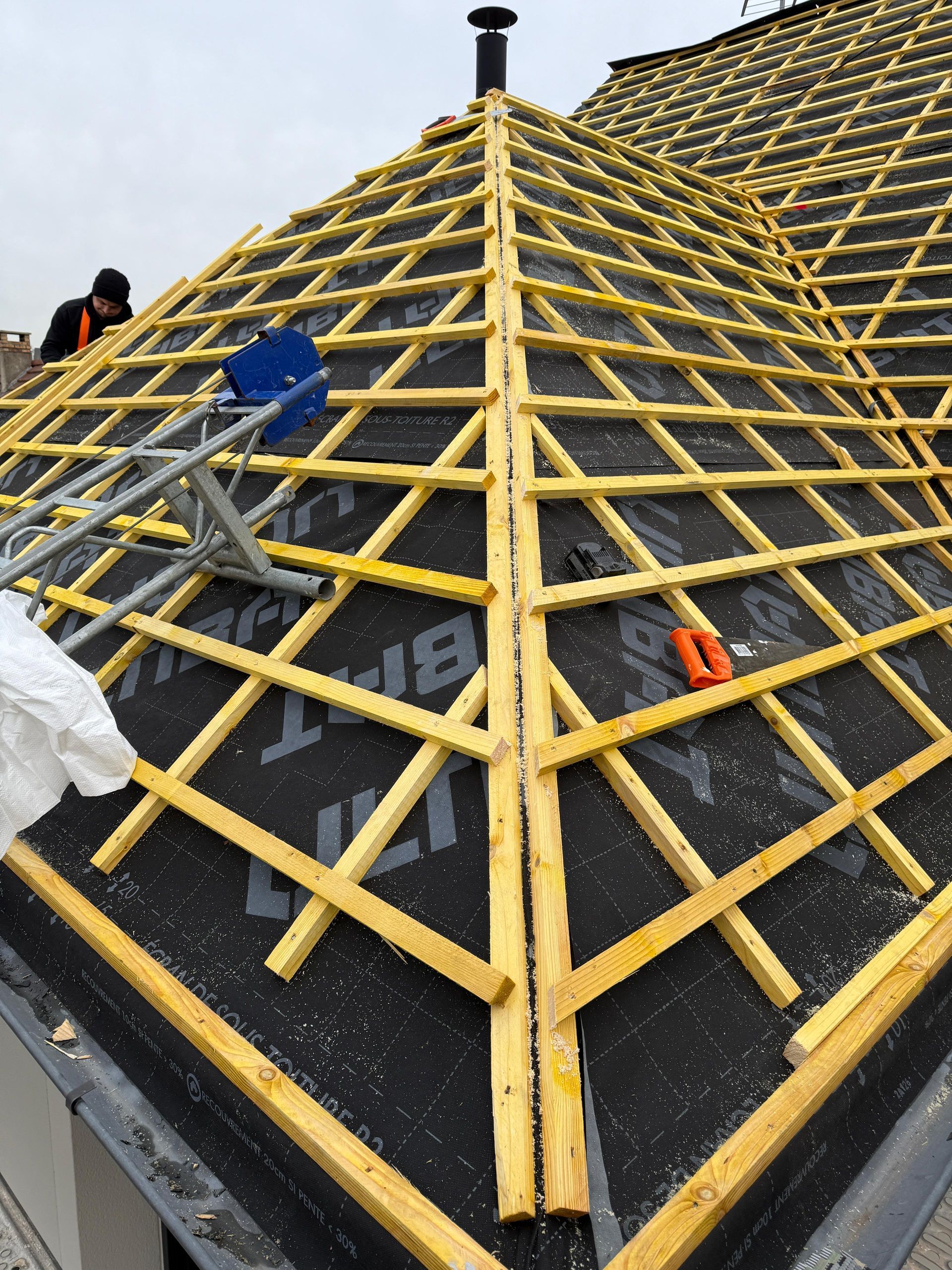 Un ouvrier installe sur le toit d'une maison une grille en bois jaune posée sur une sous-couche imperméable noire.