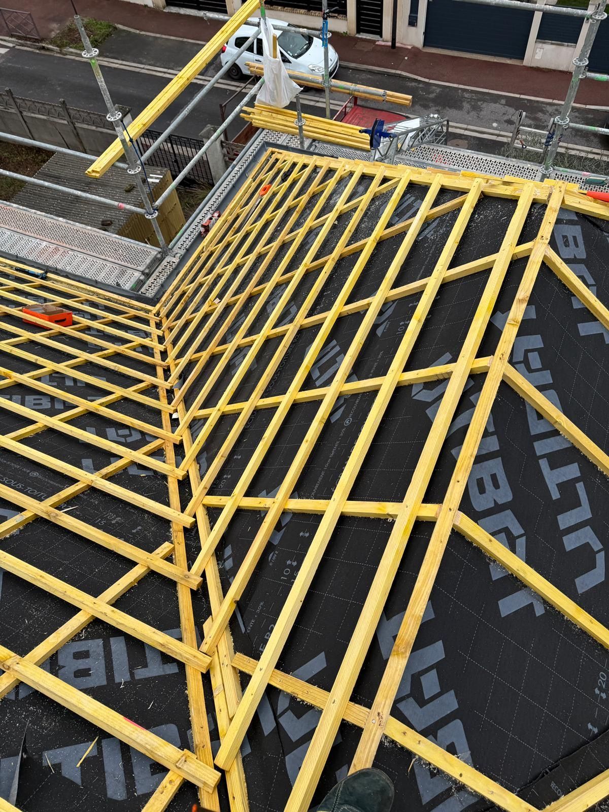 Vue aérienne d'un toit en construction avec des chevrons en bois jaune installés sur une membrane d'étanchéité noire.