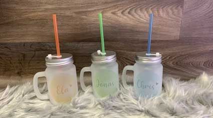 Drei Einmachglasbecher mit Strohhalmen und Namen darauf stehen auf einer Decke auf einem Holztisch.