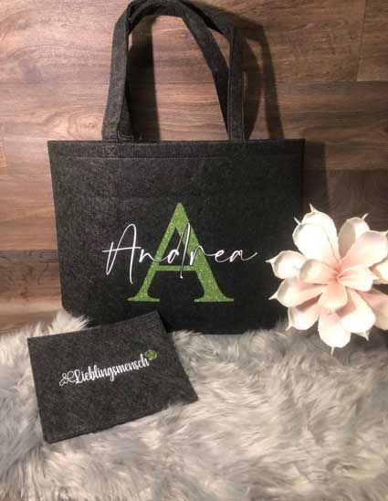 Eine schwarze Einkaufstasche mit einem grünen Buchstaben „A“ darauf liegt auf einem Holztisch neben einer Blume.