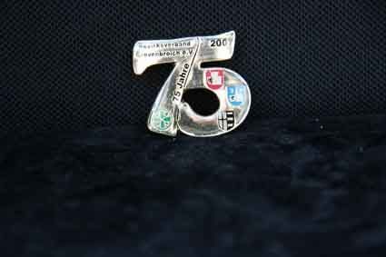 Auf einem schwarzen Untergrund sitzt eine silberne Brosche in Form der Zahl 75.