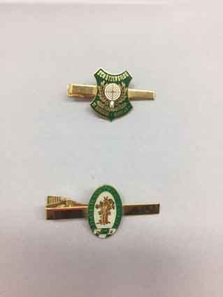 Auf einer weißen Fläche liegen zwei Krawattenklammern mit Emblemen darauf.