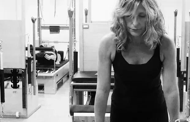 Mujer con vestido negro de pie en un gimnasio, inclinada hacia adelante con pesas y equipo detrás de ella.