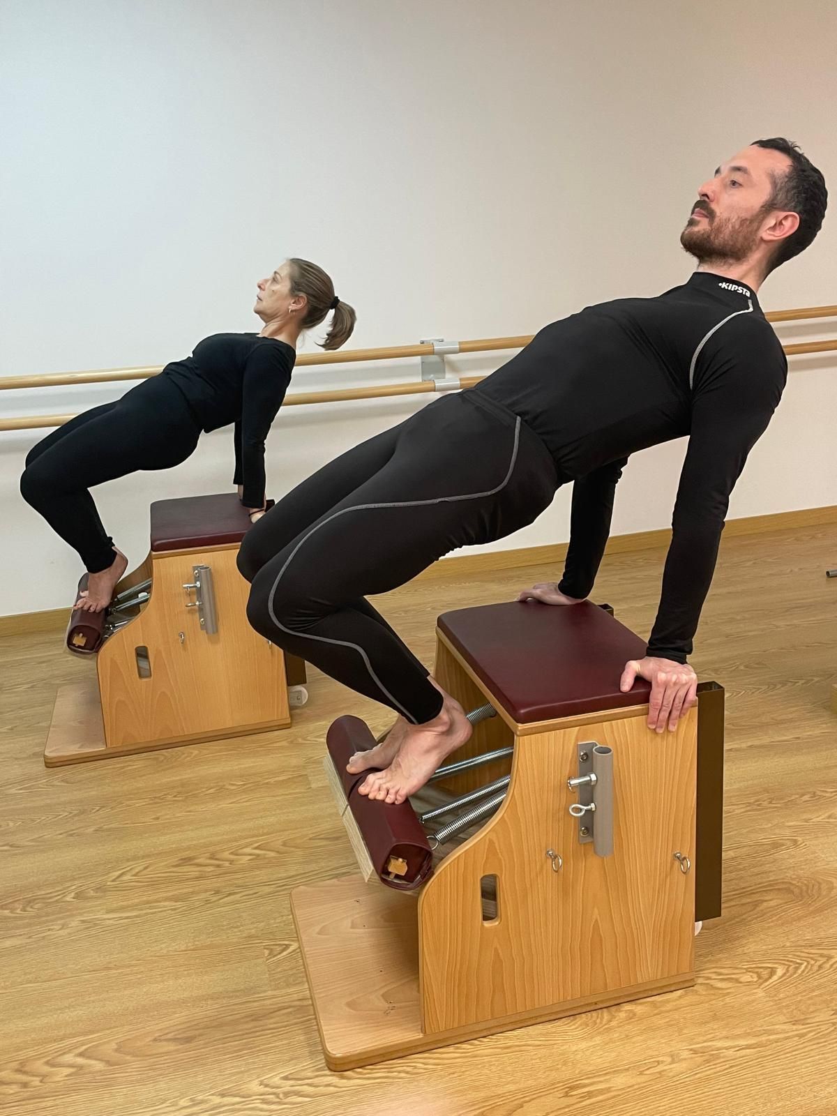 Dos personas haciendo la postura del puente en sillas de yoga de madera en un estudio.