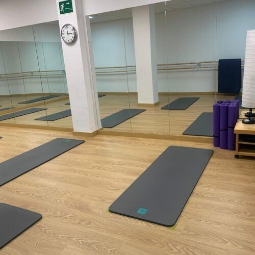 Estudio de yoga vacío con espejos, esterillas grises sobre suelo de madera y rodillos de espuma morados junto a la pared.