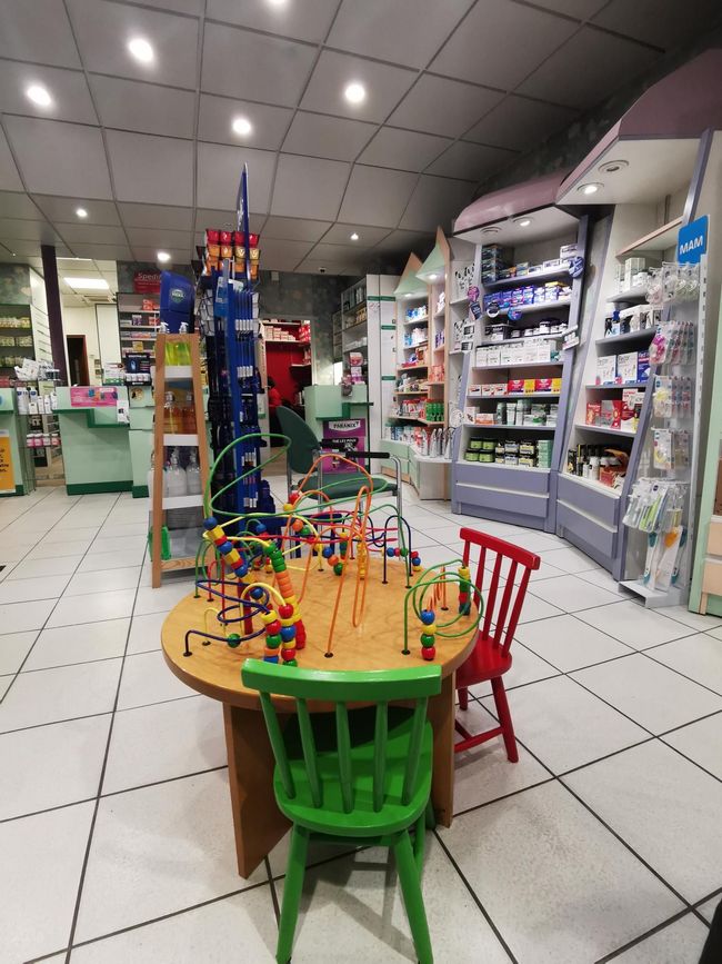 Intérieur d'une pharmacie avec un espace de jeux pour enfants. Une table en bois avec des jouets et des chaises colorées. Des étagères remplies de produits.