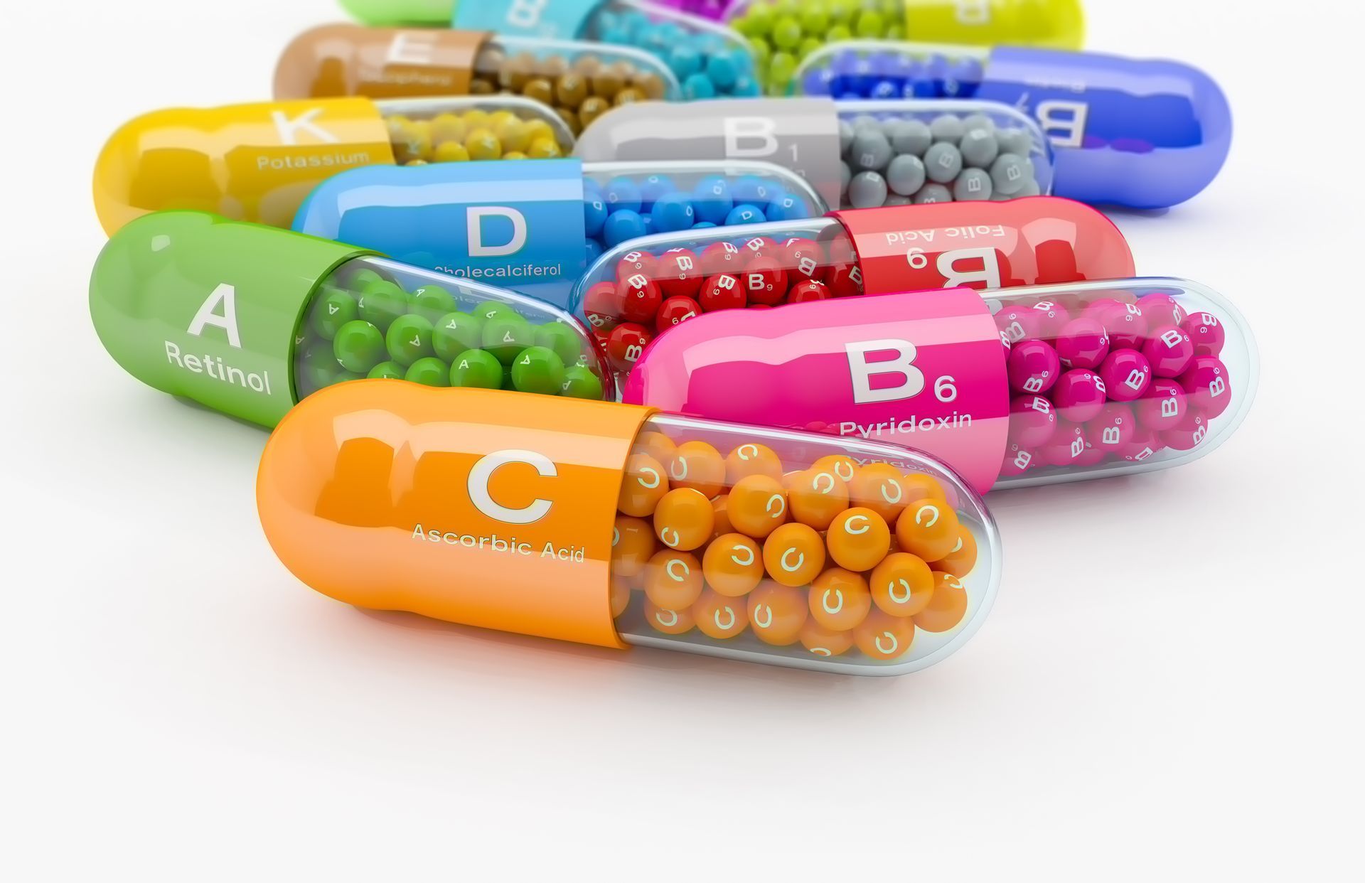Assortiment de capsules vitaminées colorées, étiquetées de A à K, disposées sur une surface blanche.