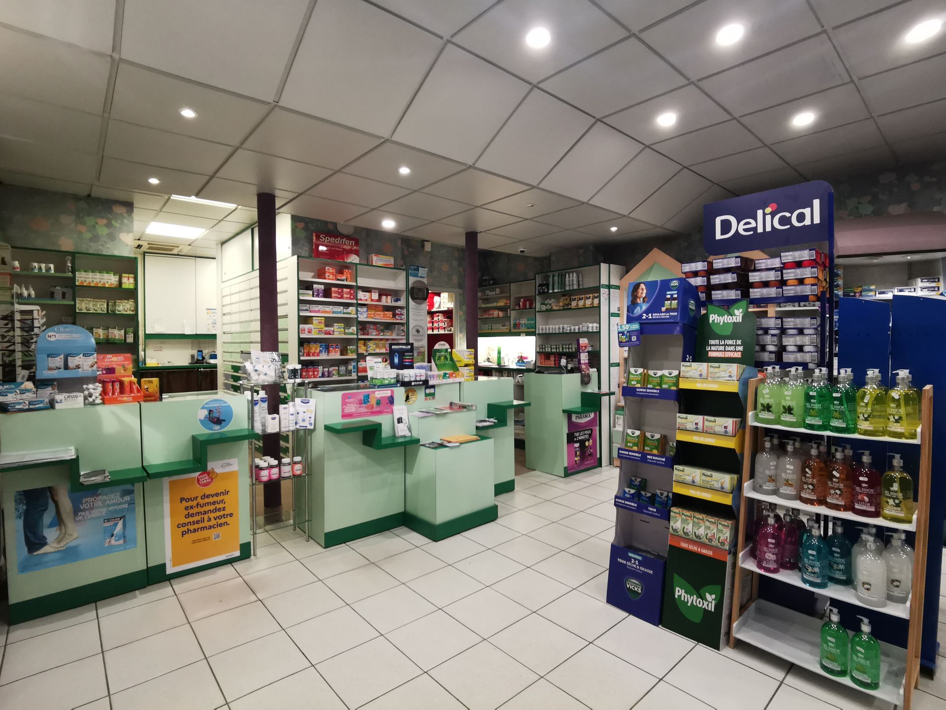 Intérieur d'une pharmacie avec des comptoirs vert clair, un sol carrelé blanc et diverses étagères remplies de produits de santé.