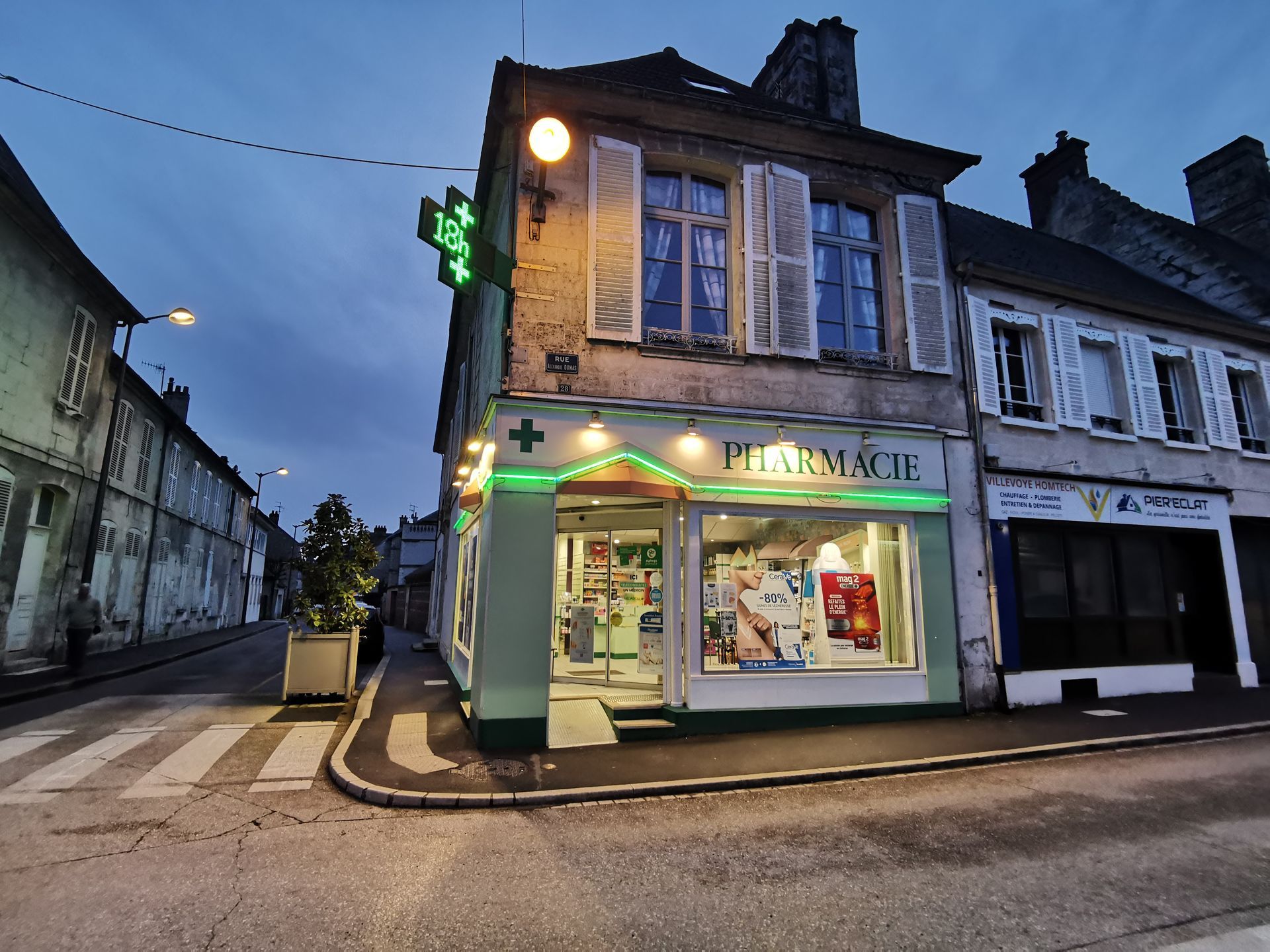 La façade d'une pharmacie d'angle en France, illuminée par une croix verte au crépuscule, avec des bâtiments adjacents à proximité.