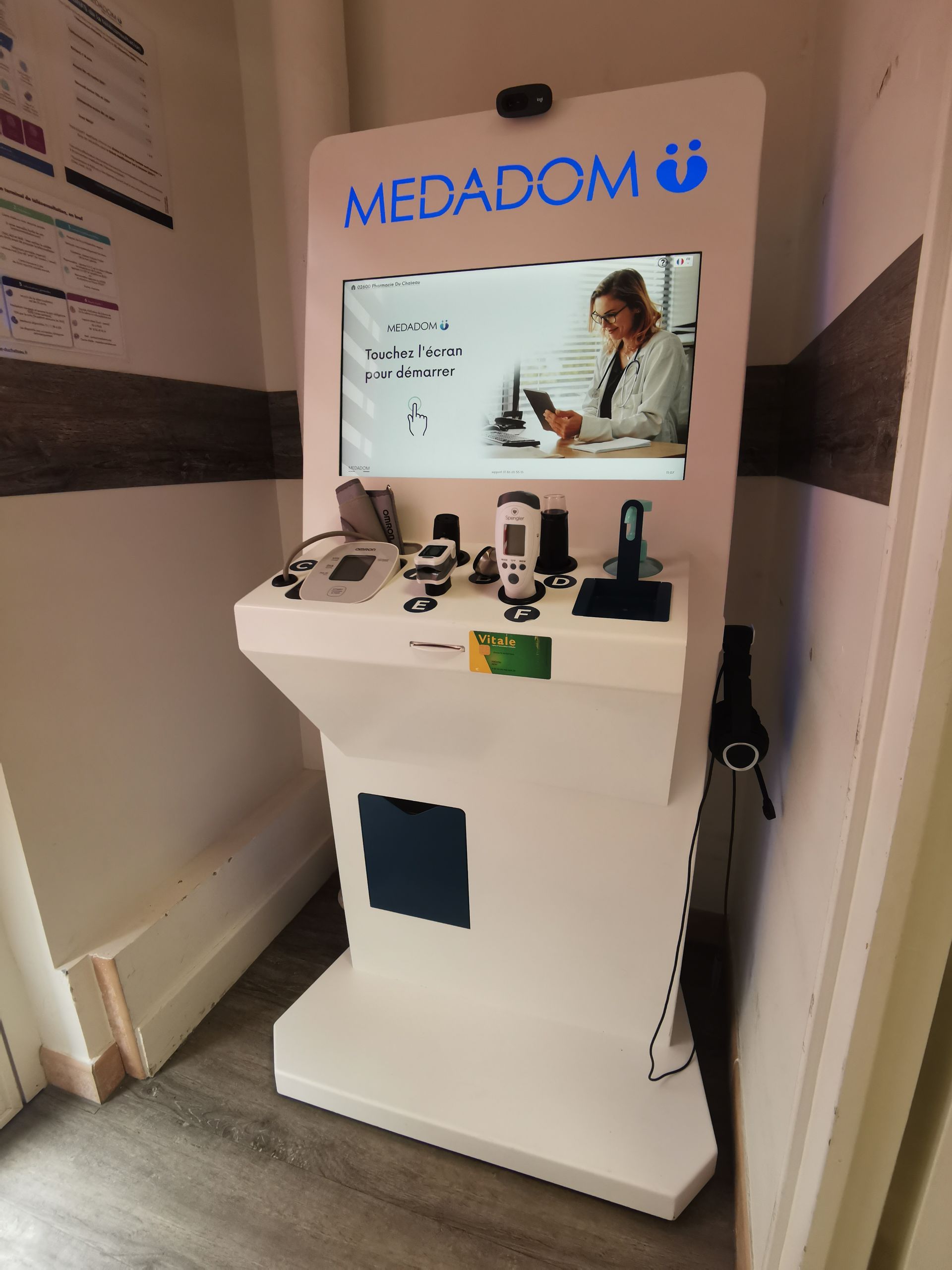 Borne de télémédecine Medadom blanche avec écran, dispositifs médicaux intégrés et caméra, située dans un espace clinique.
