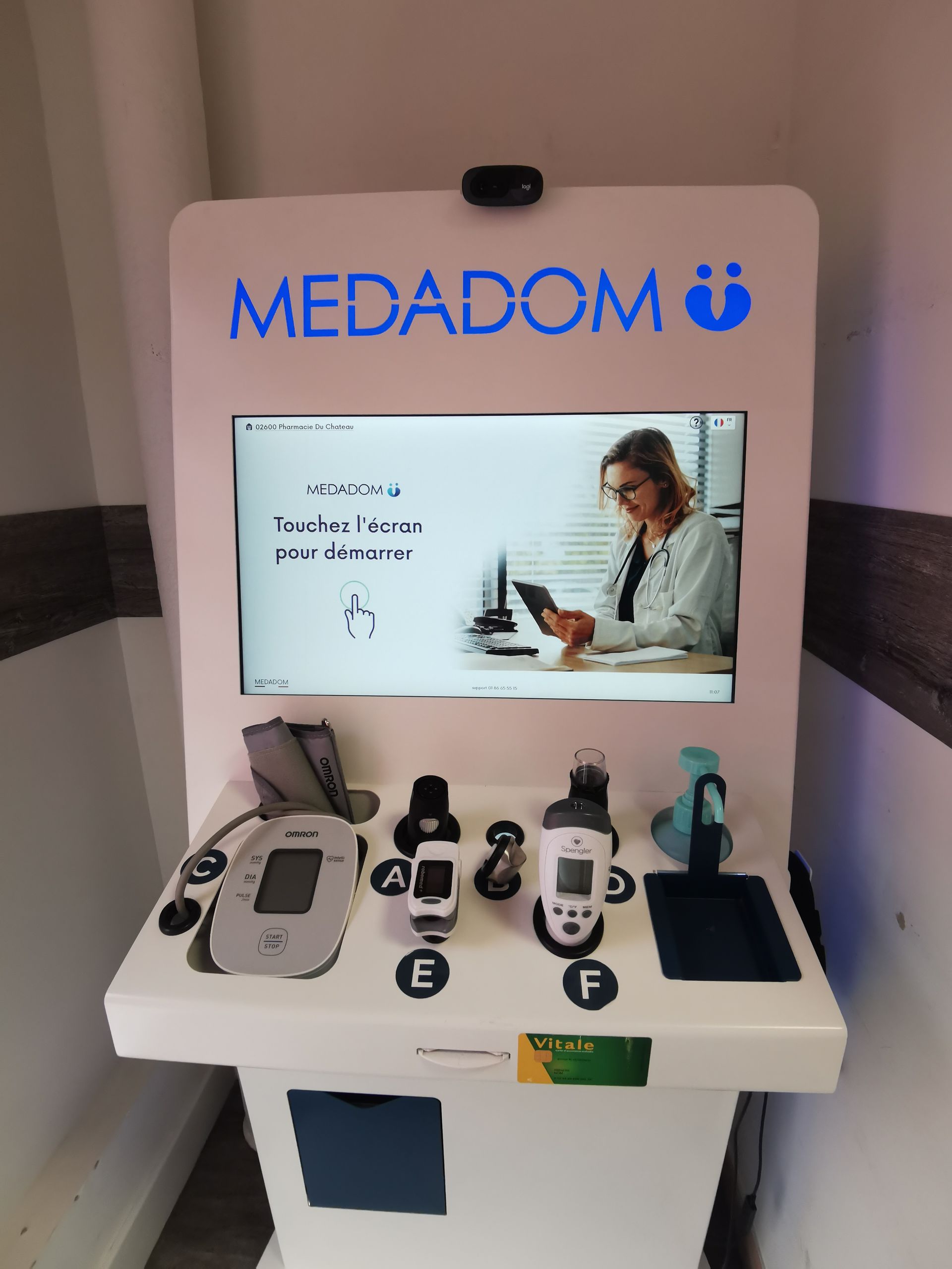 Une borne de télémédecine Medadom avec un écran, des outils de diagnostic médical et des étiquettes de A à F, placée contre un mur.