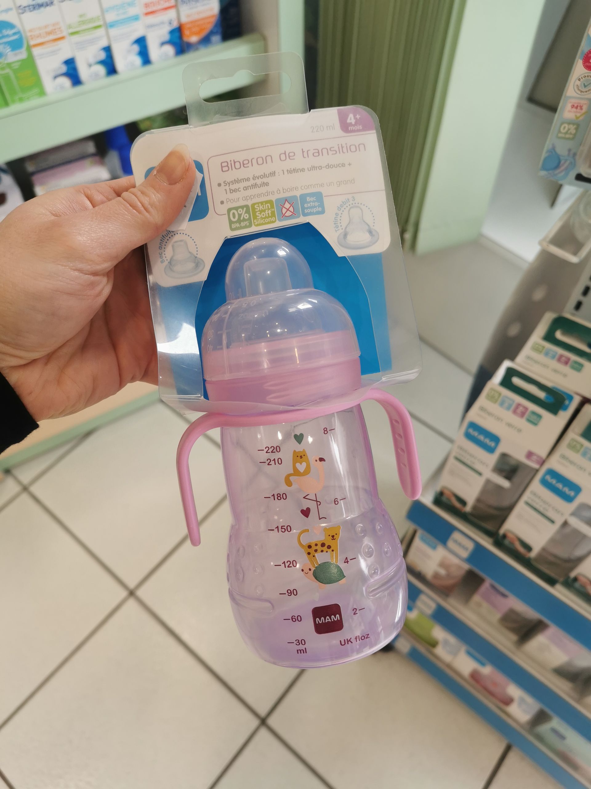 Dans un magasin, une main tient un biberon rose pour bébé de la marque MAM, avec des poignées et des illustrations d'animaux.