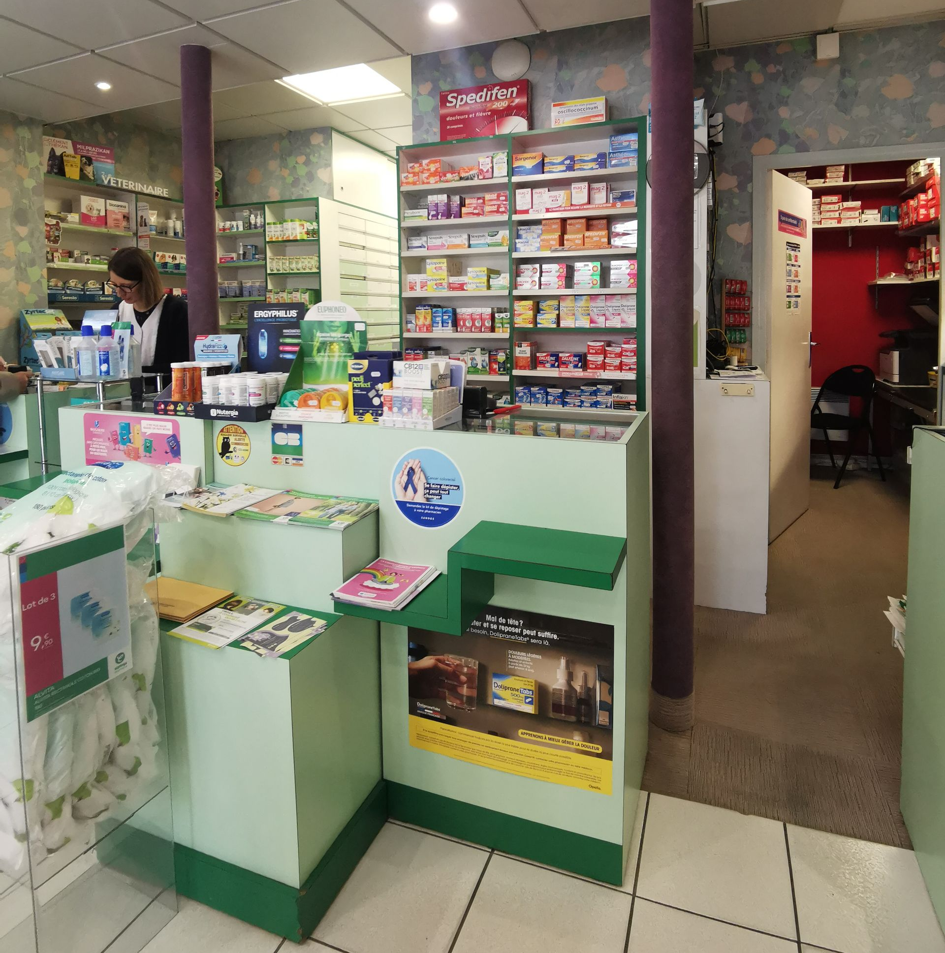 Comptoir de pharmacie avec un membre du personnel, étagères de produits médicaux et porte donnant sur une zone de stockage.