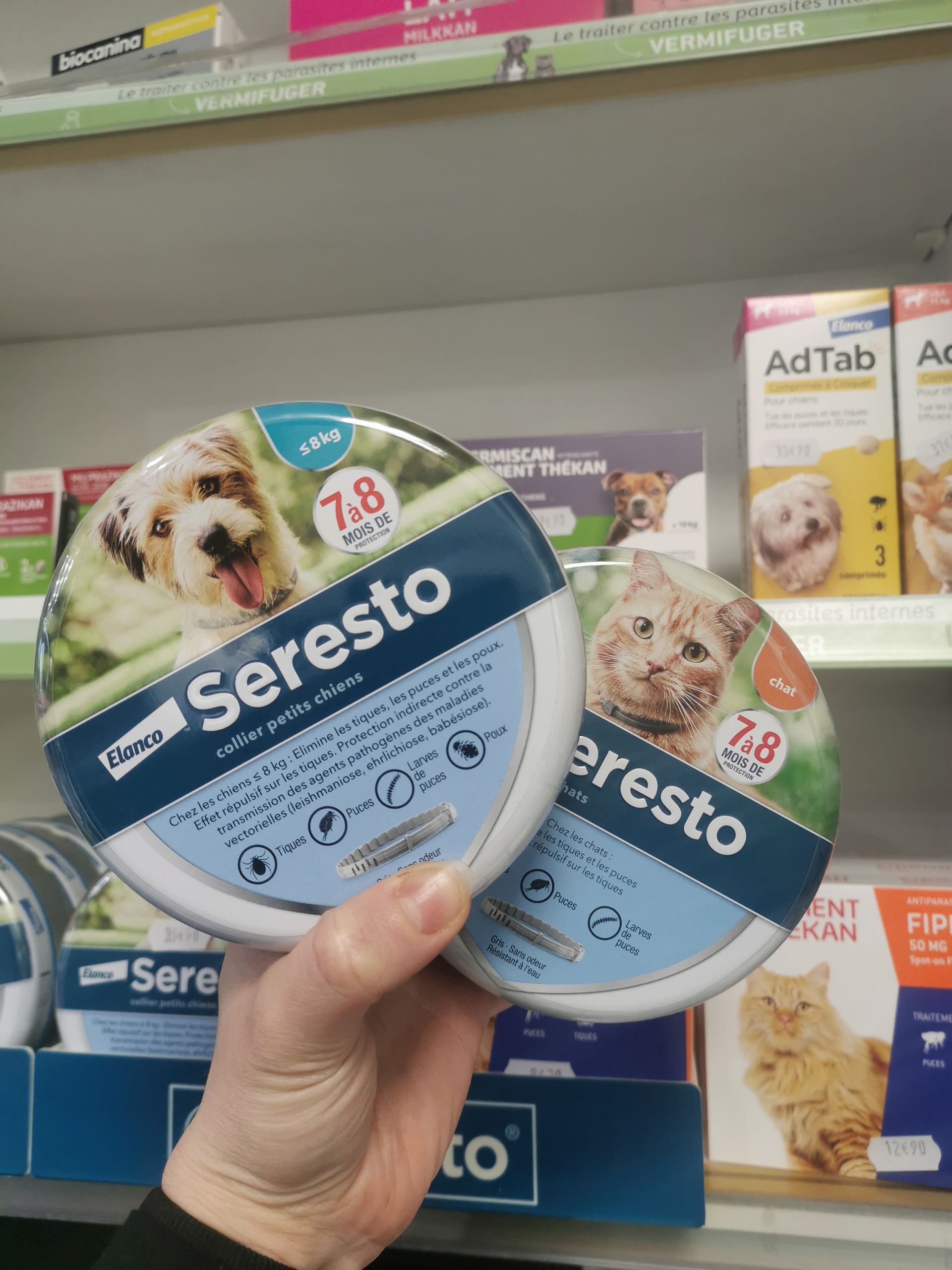 Dans un magasin, une main tient deux emballages ronds de colliers anti-puces et anti-tiques Seresto, un pour chiens et un pour chats.