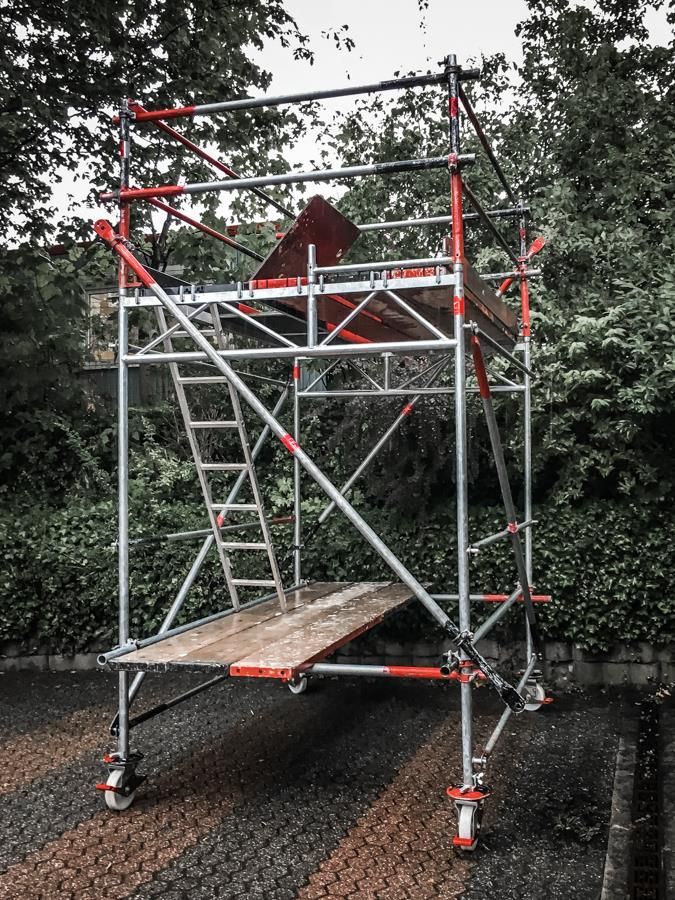 Auf einem Gehweg steht ein Gerüstturm mit einer Holzplattform und Treppen.