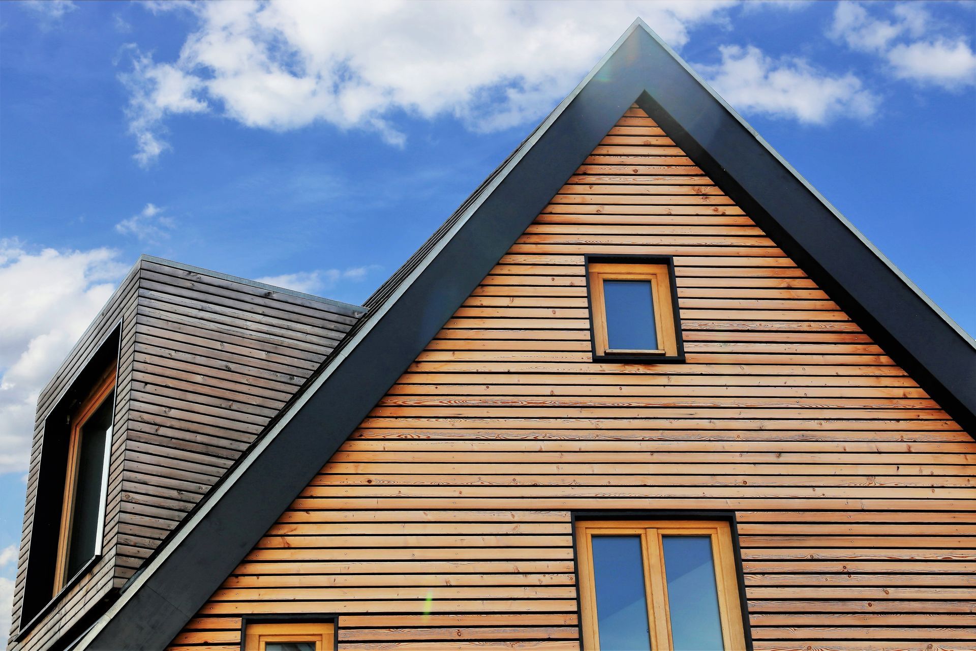 Maison moderne à pignon avec bardage en bois, boiseries foncées et ciel nuageux.