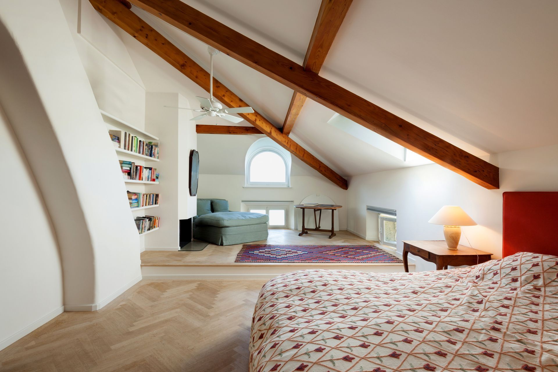 Chambre mansardée lumineuse avec poutres apparentes, un lit, un fauteuil, une bibliothèque et une fenêtre cintrée.