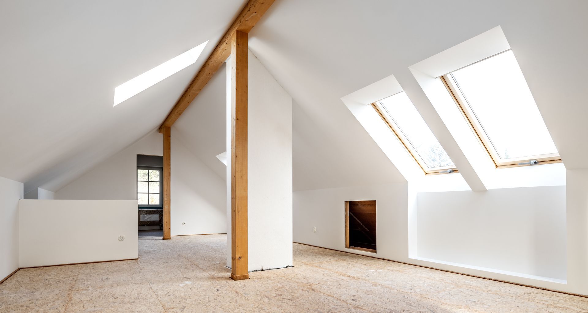 Pièce mansardée vide avec murs blancs, poutres apparentes, plafond mansardé et fenêtres de toit.