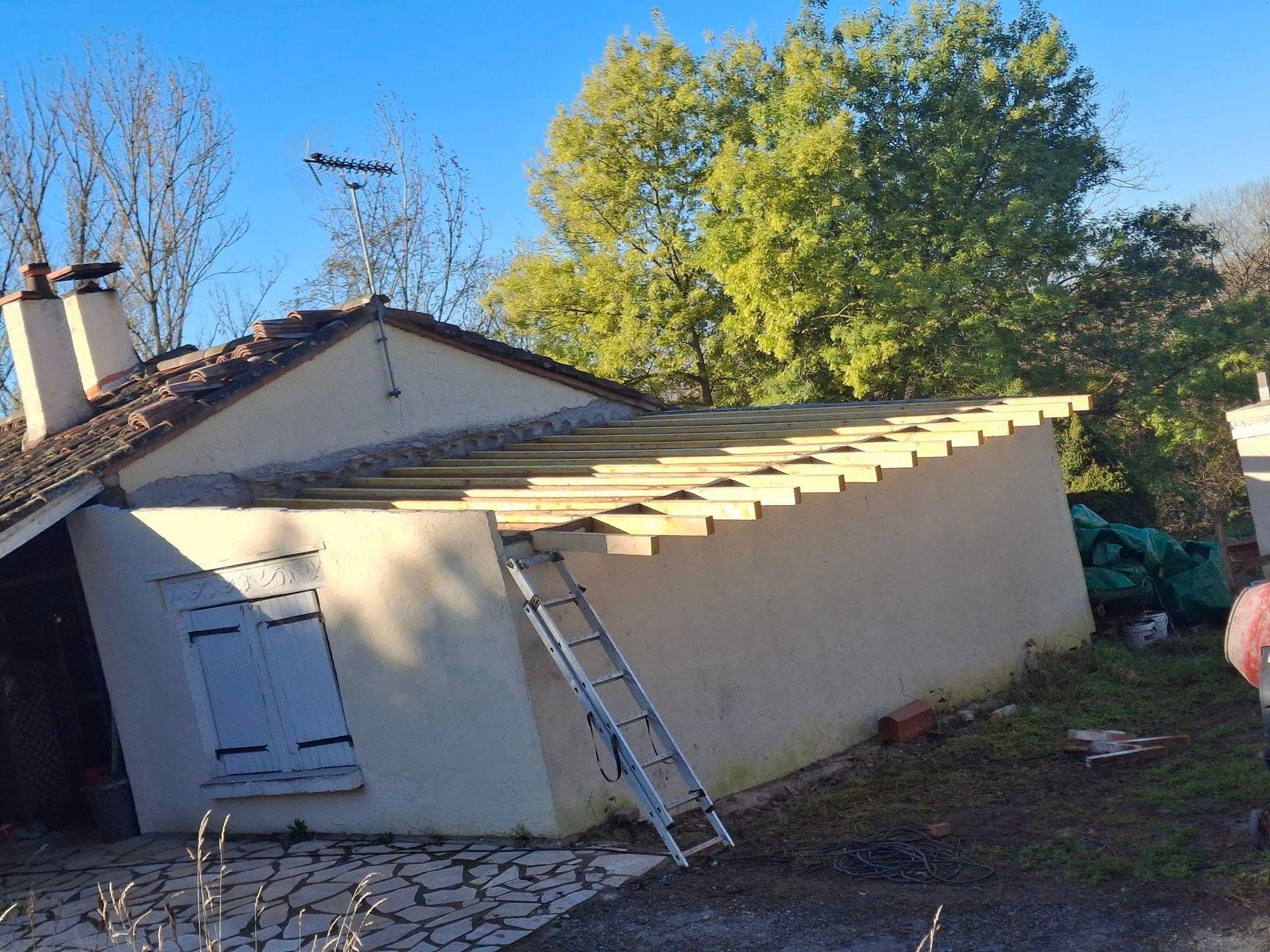 Extension d'une maison en construction, avec une échelle appuyée contre le mur et des arbres en arrière-plan.