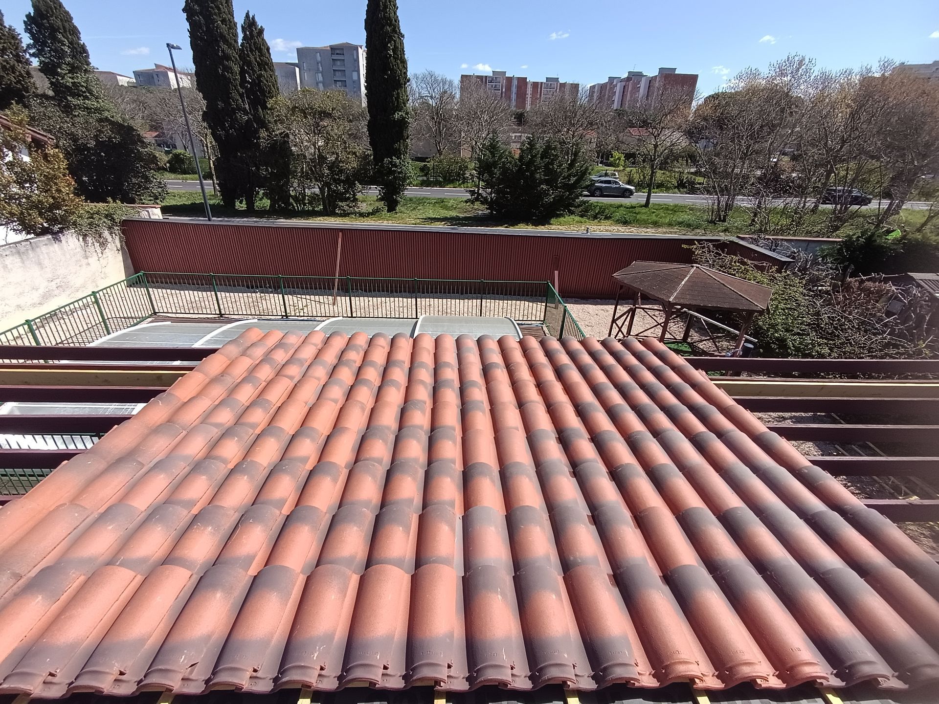 Toit de tuiles en terre cuite au premier plan, jardin et immeubles en arrière-plan.