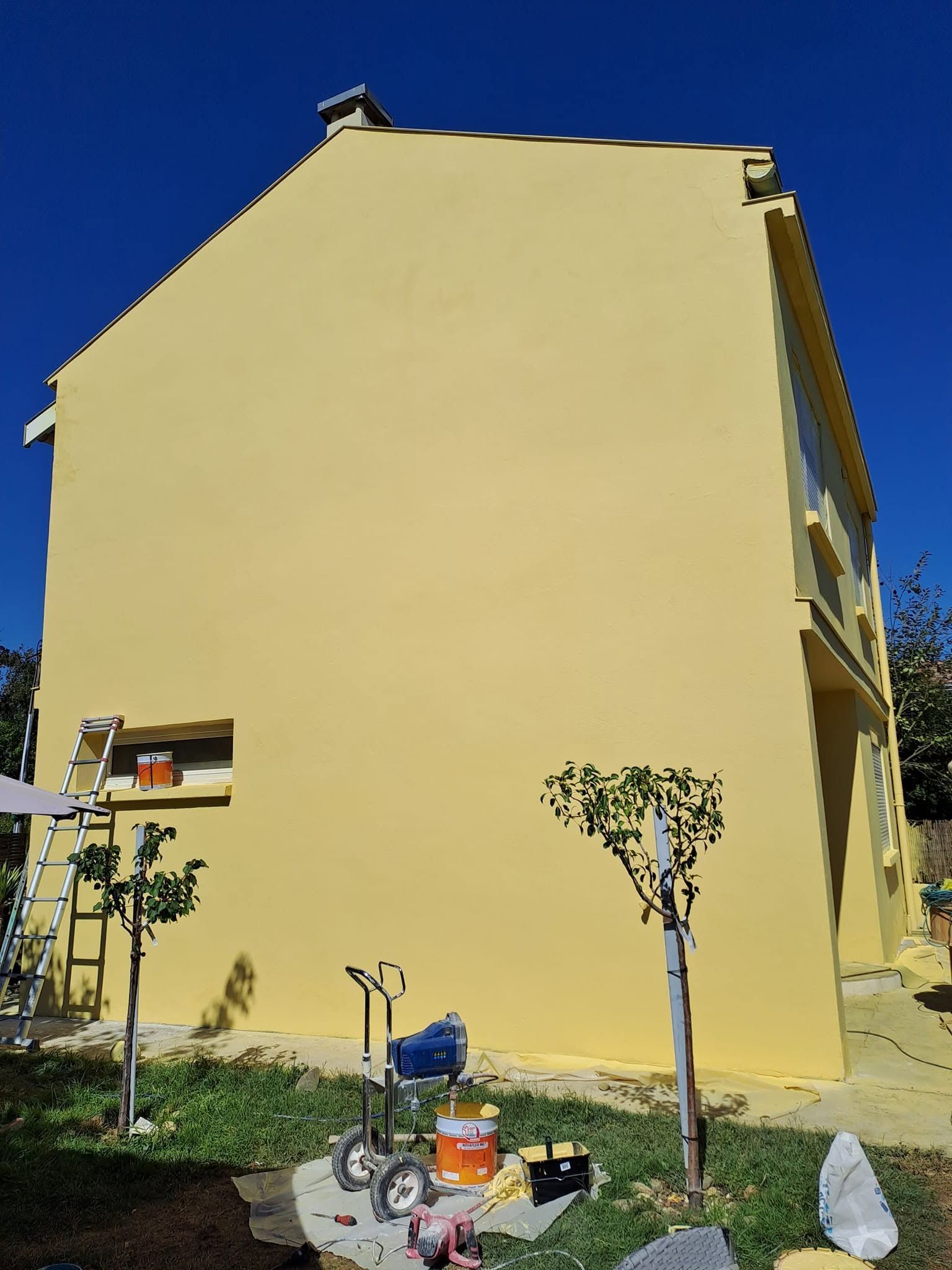 Mur jaune d'une maison sous un ciel bleu avec de petits arbres et un vélo au premier plan.