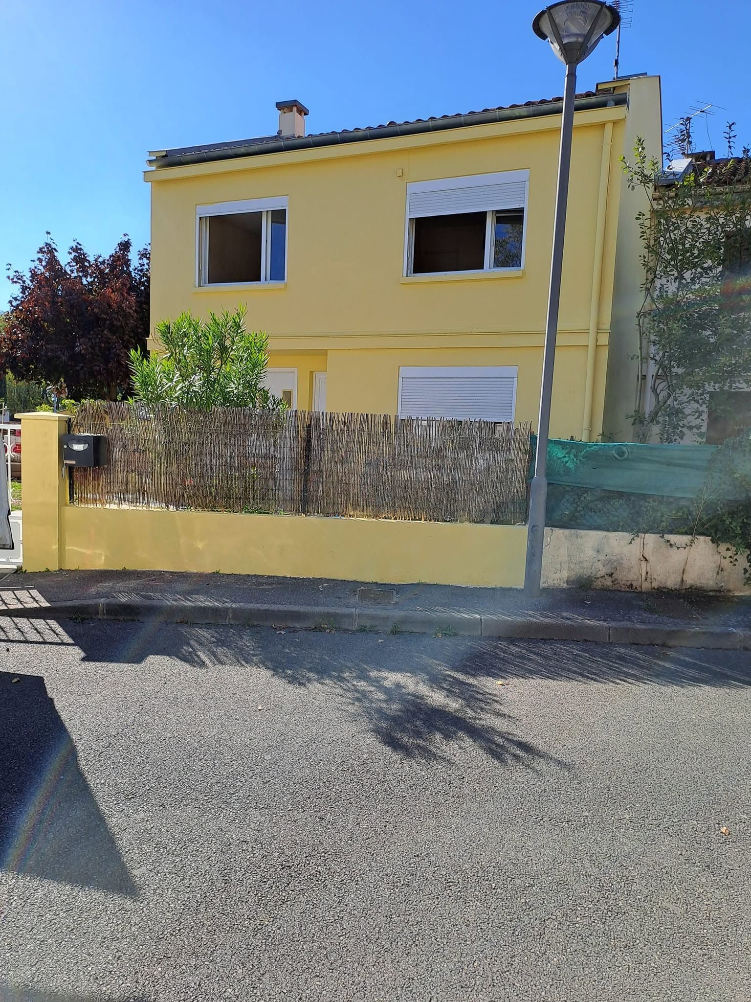 Maison jaune à deux étages, volets blancs, cour clôturée et lampadaire en premier plan.