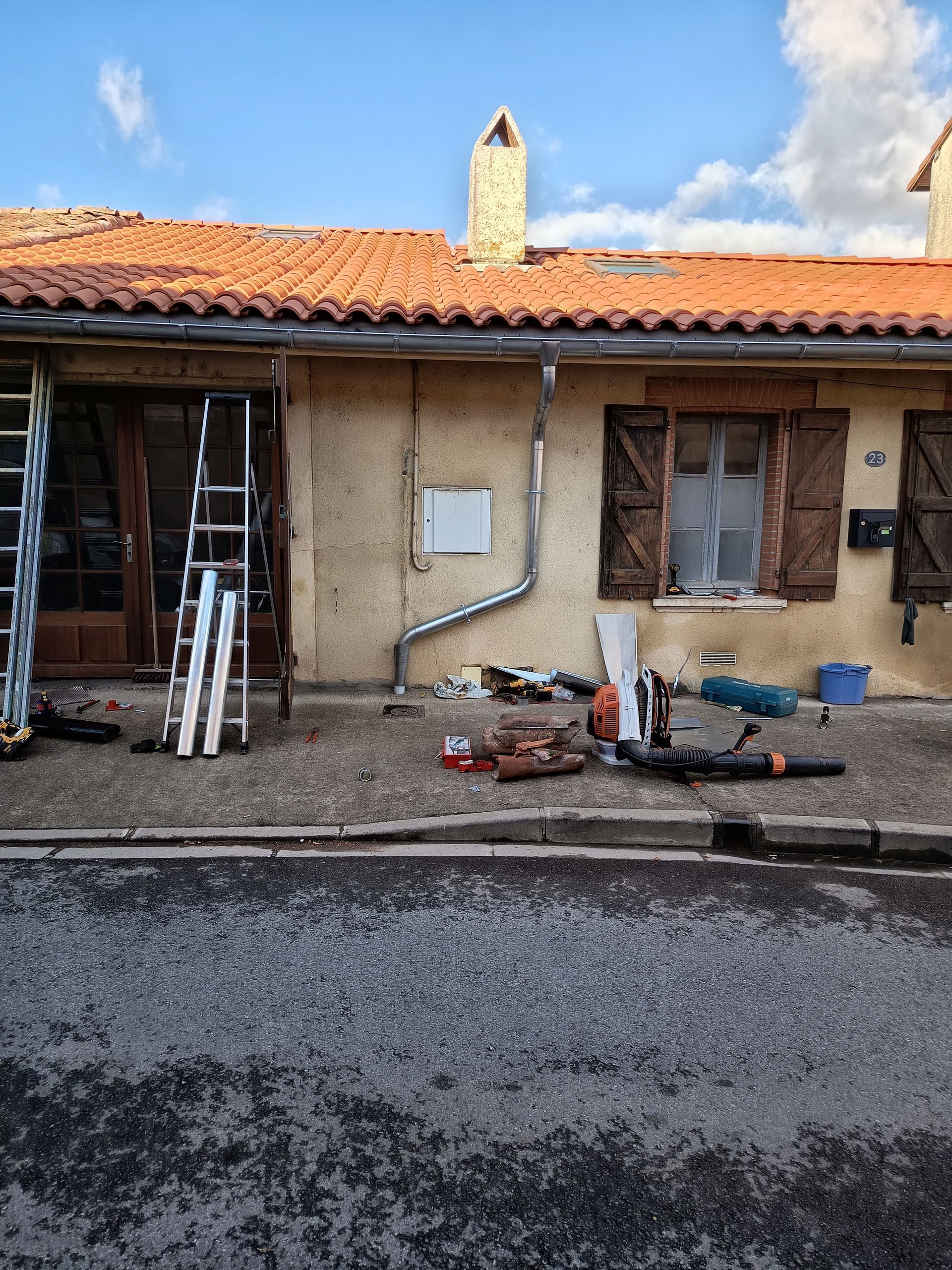 Vue de face d'une maison en stuc avec un toit de tuiles, des outils et des matériaux au sol, et une porte entrouverte.
