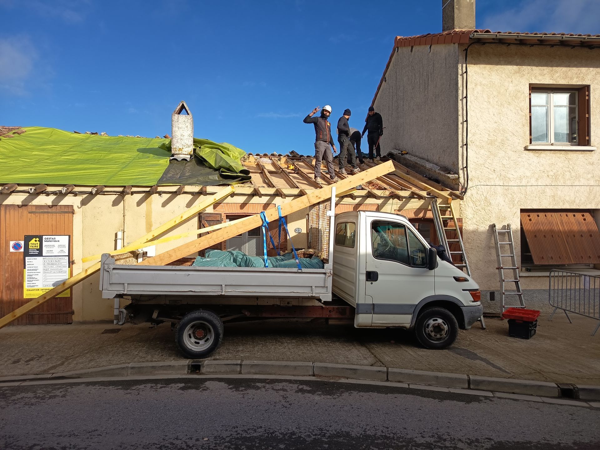 Des ouvriers réparent un toit à côté d'un camion plateau blanc dans une rue, des débris et des outils sont visibles.