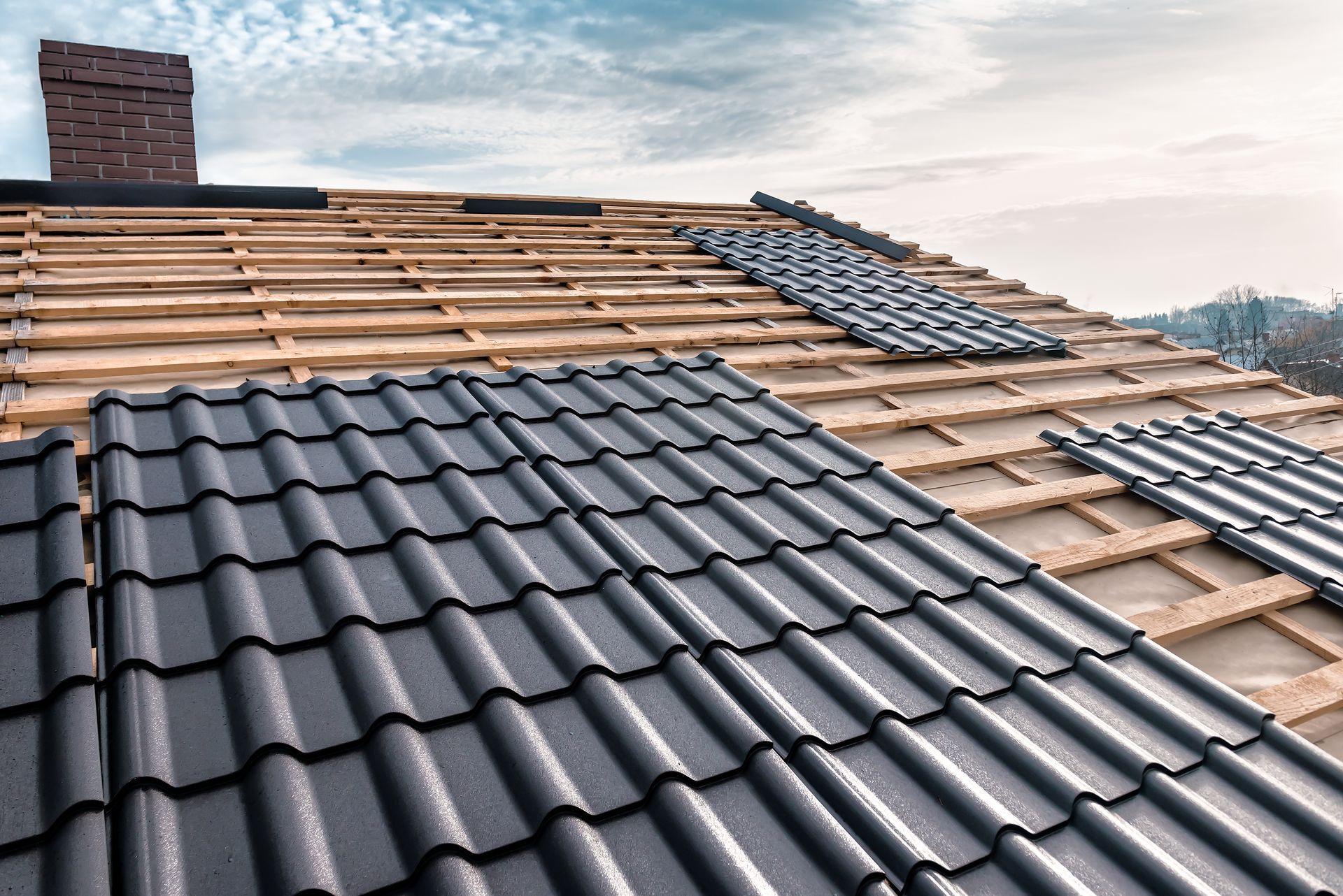 Toiture en construction avec des tuiles grises et une cheminée en briques sous un ciel nuageux.