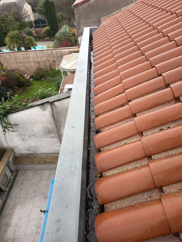 Vue d'un toit de tuiles en terre cuite rouge et d'une gouttière blanche donnant sur un jardin.