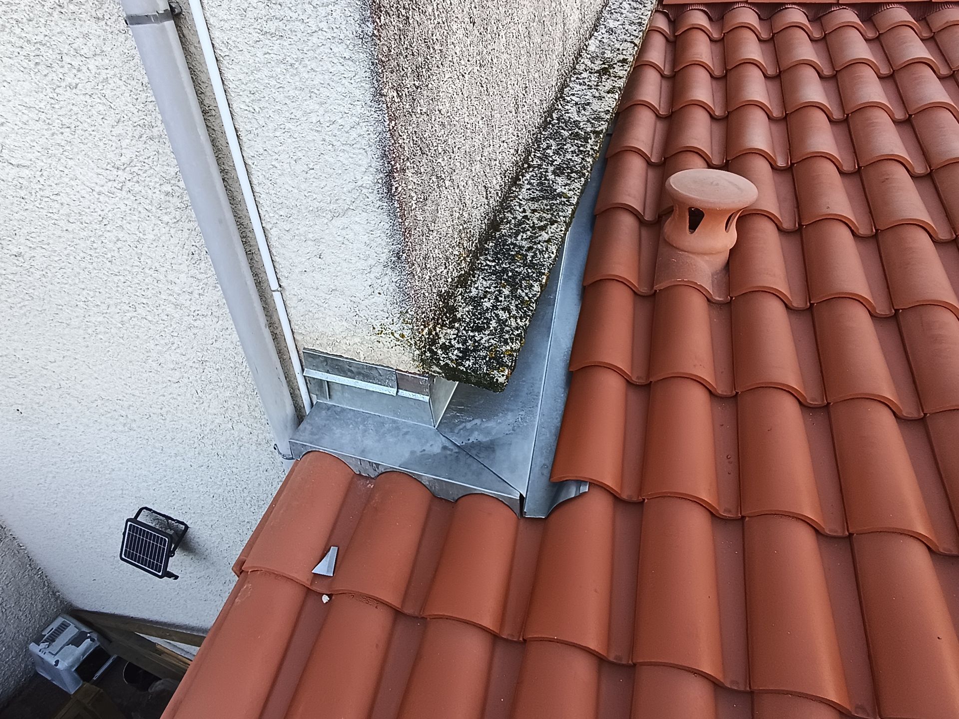 Toit de tuiles rouges jouxtant un mur blanc, avec une bande en zinc autour d'un joint d'angle.