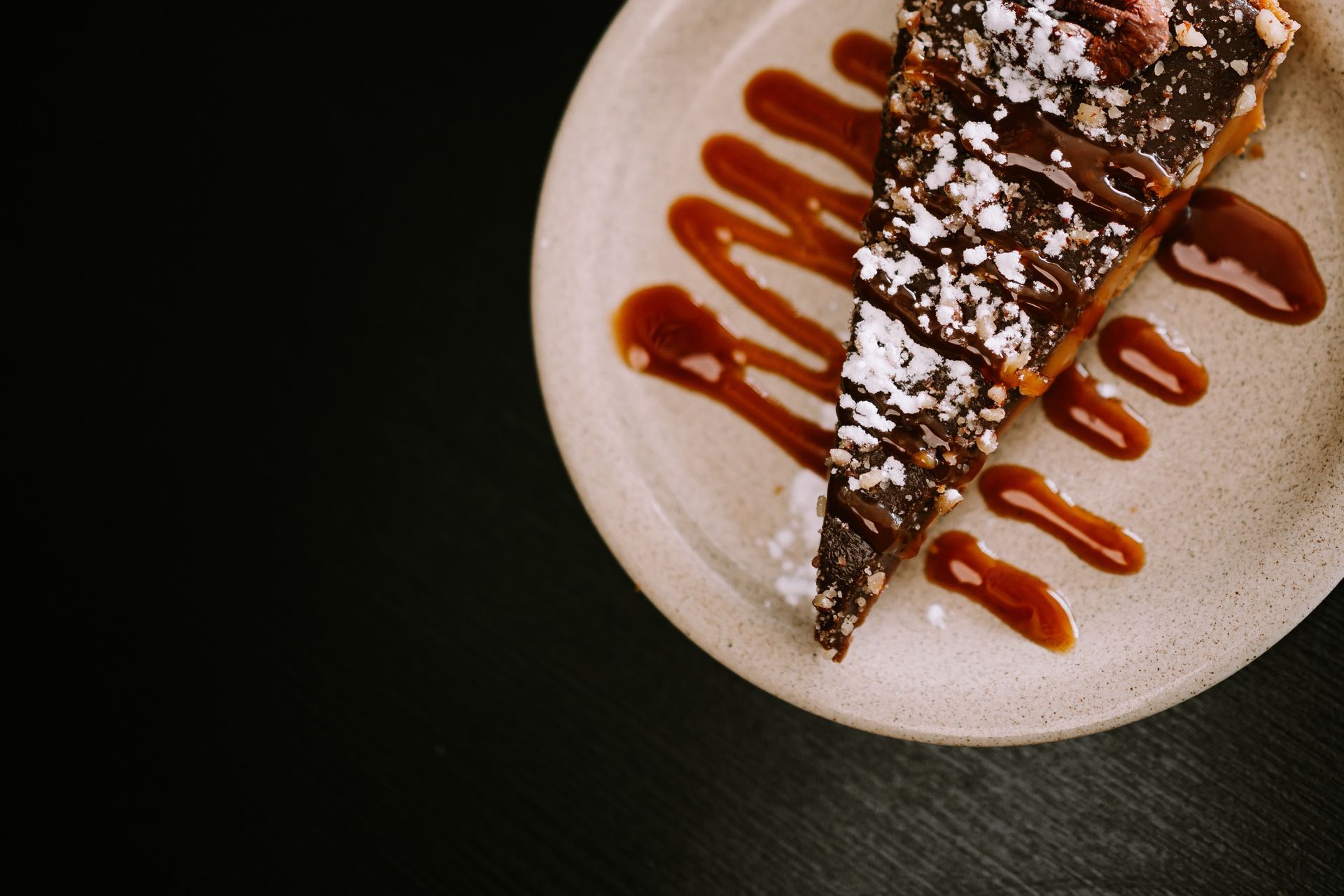 Fatia de bolo de sobremesa com calda de caramelo e açúcar de confeiteiro em um prato contra um fundo escuro.