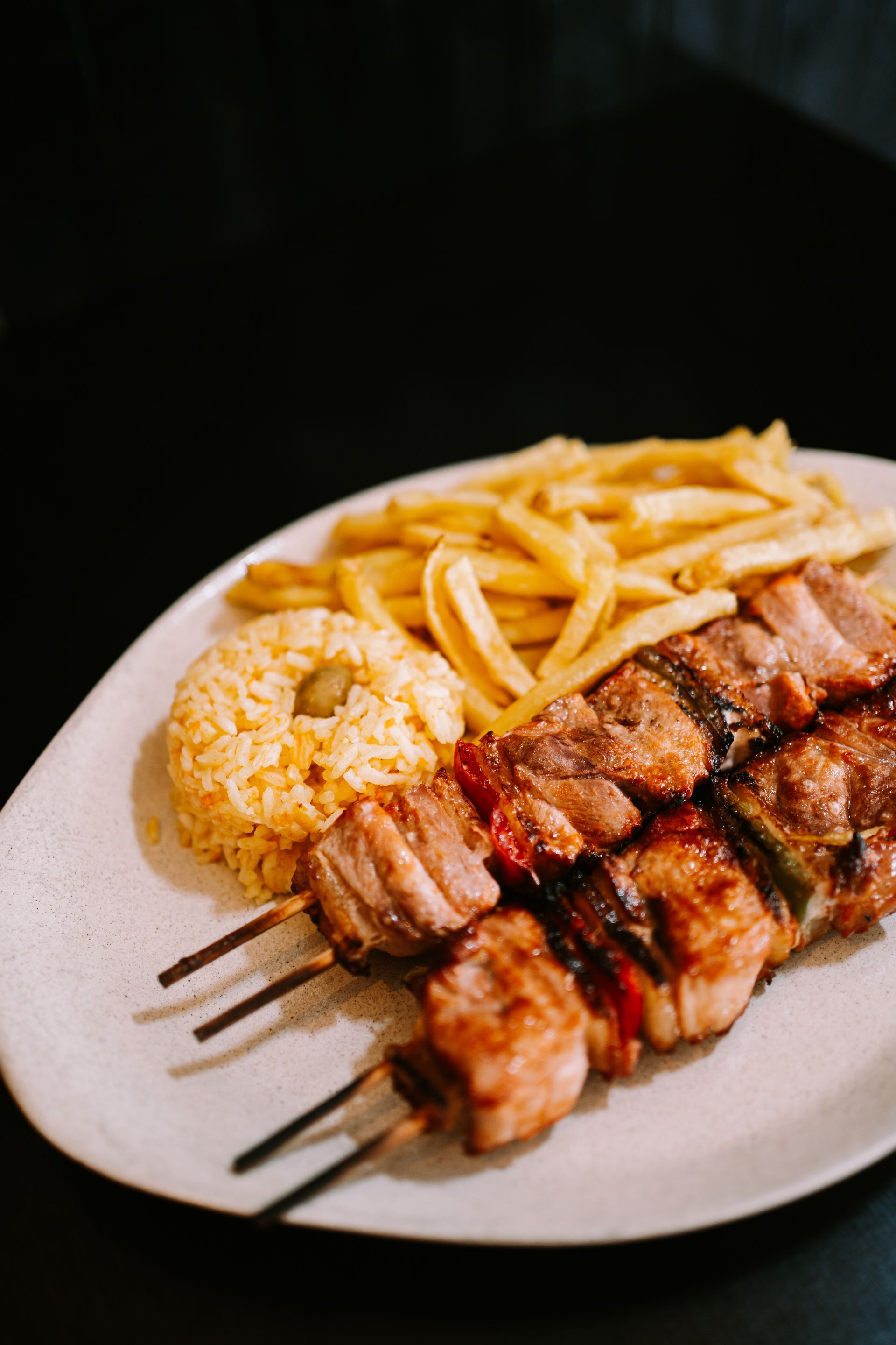 Espetinhos de carne, batatas fritas e arroz em um prato branco sobre um fundo preto.