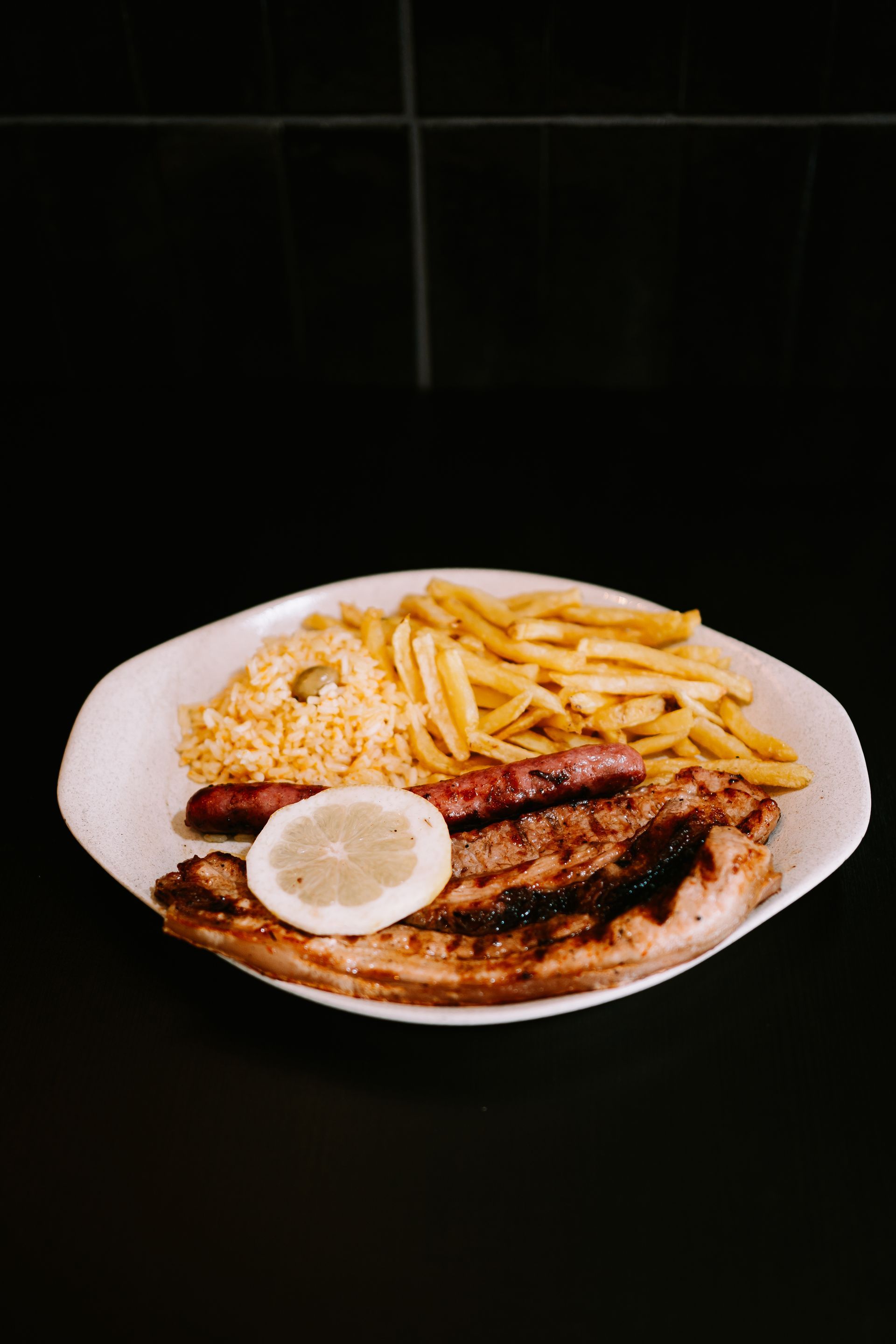 Carne grelhada, linguiça, batatas fritas e arroz em um prato branco, com uma fatia de limão. Tudo sobre um fundo preto.