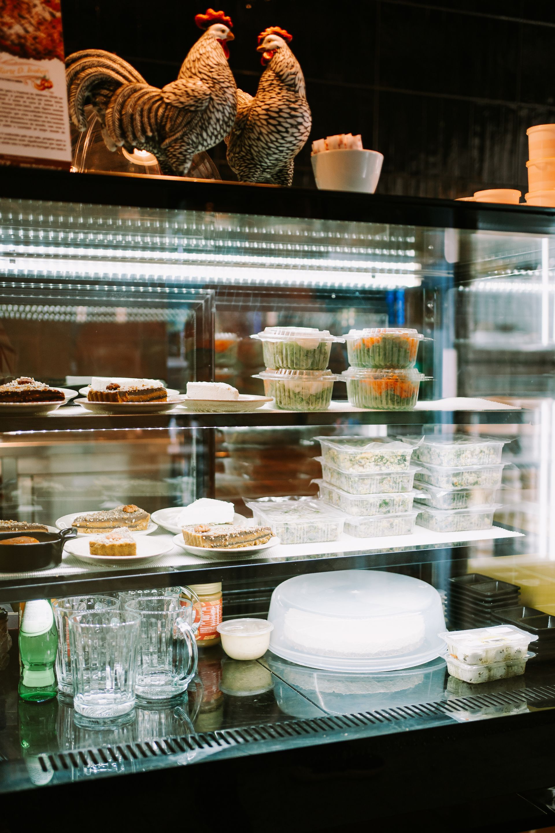 Vitrine com doces, estátuas de galo e bebidas em um ambiente de café.
