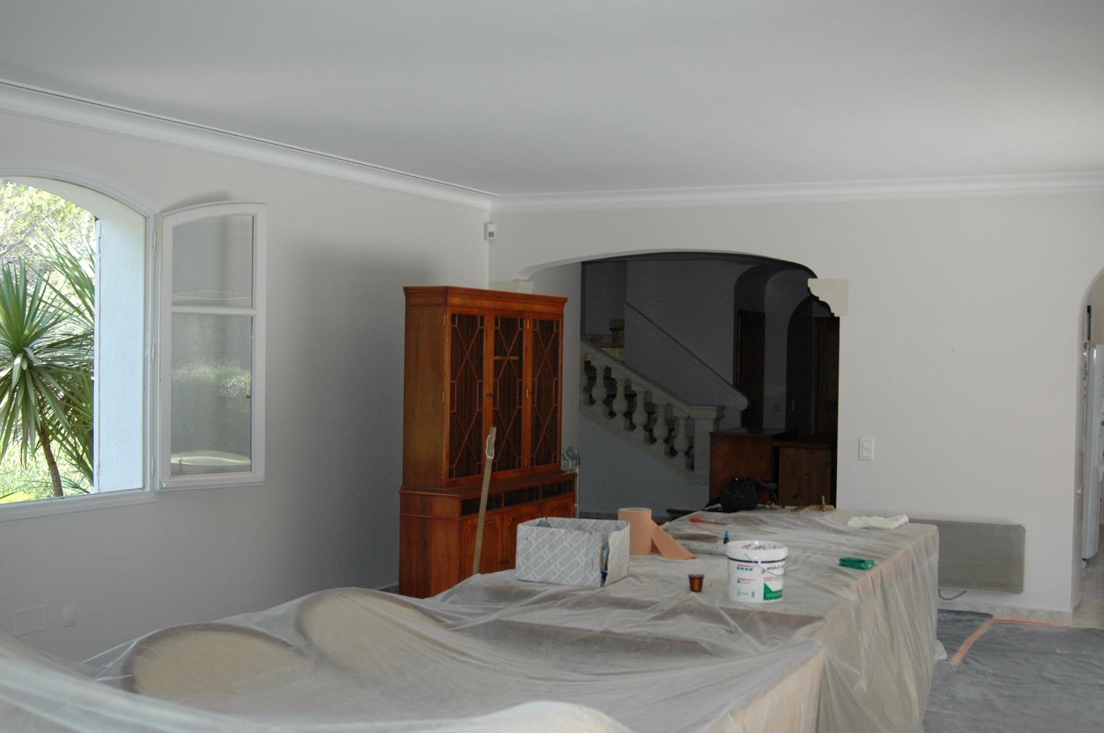 Salon avec meubles recouverts de tissu, murs blancs, armoire en bois et porte cintrée donnant sur l'escalier.