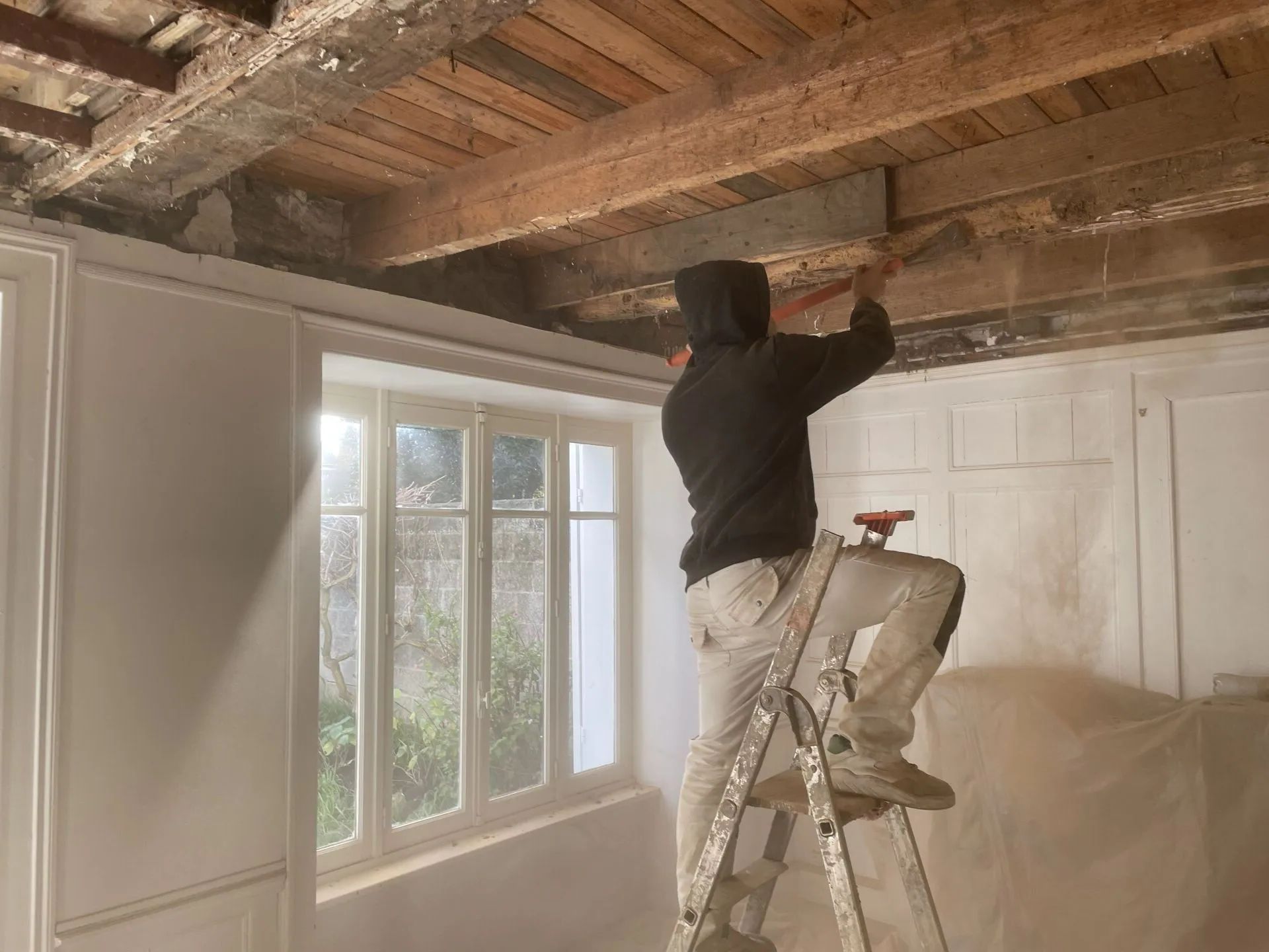Une personne sur un escabeau traite les poutres apparentes d'un plafond en bois.