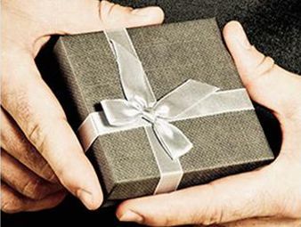 Manos sosteniendo una pequeña caja de regalo envuelta en papel gris con una cinta y un lazo plateados.